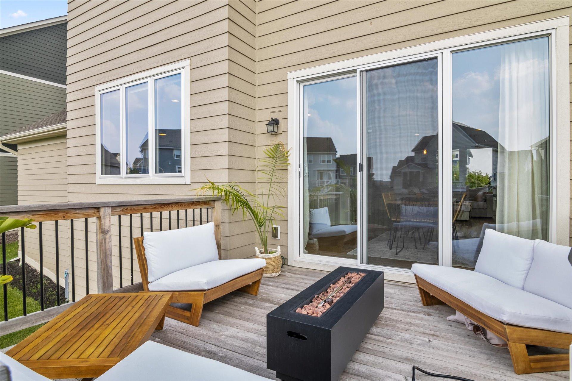 Wooden deck with cushioned lounge chairs and a black fire table beside sliding glass doors.