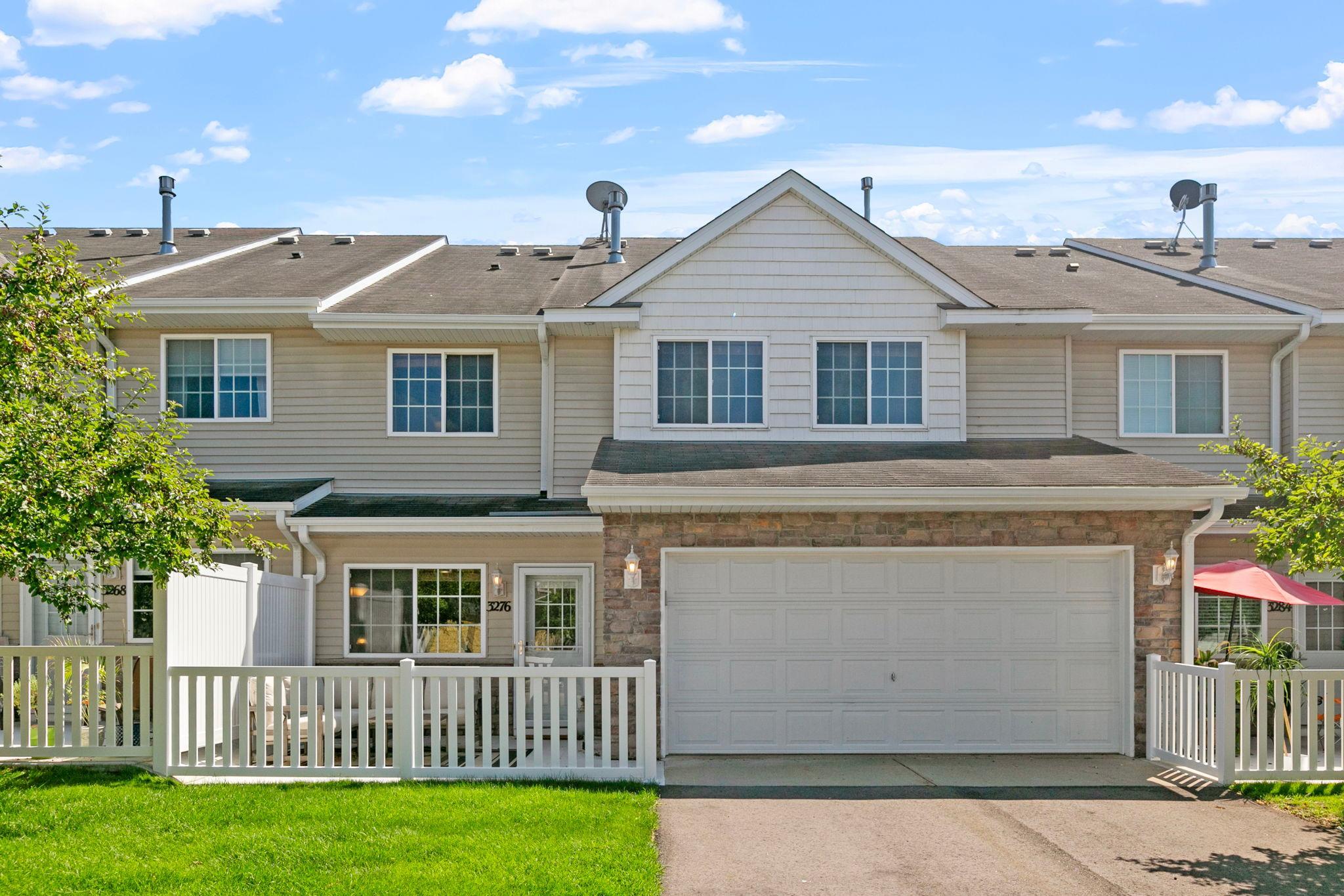 Two-story townhouse with a stone-front garage, white fencing, and a green lawn.
