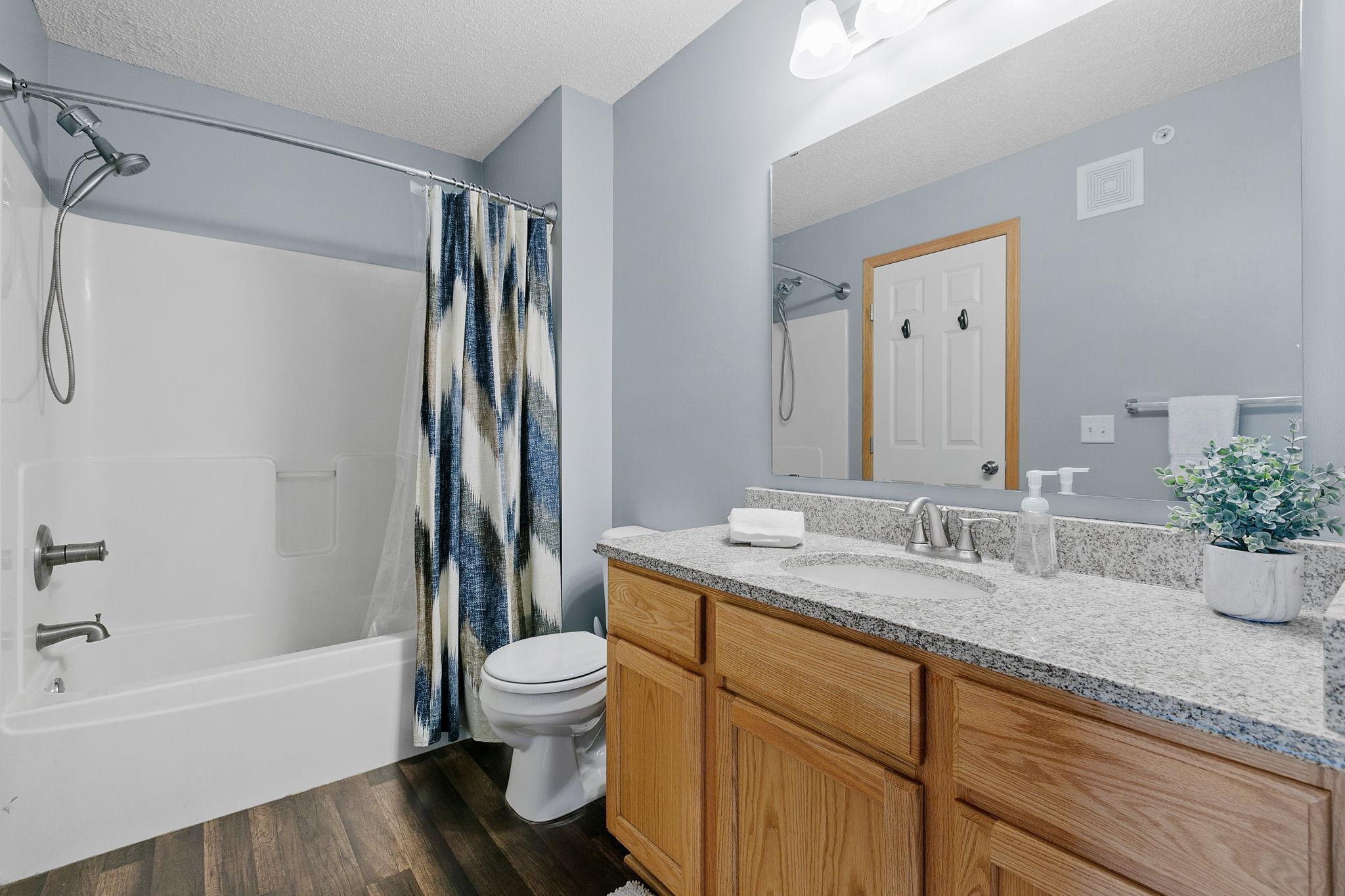 Bathroom with a white tub, patterned shower curtain, and oak vanity with a speckled countertop.