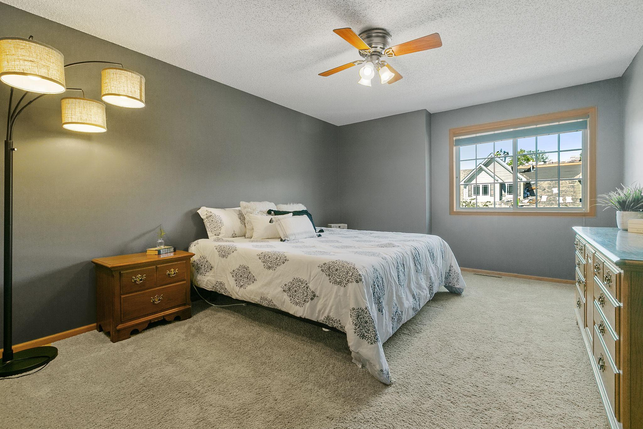 Furnished bedroom with gray walls, patterned white bedding, wooden furniture, and a large window.