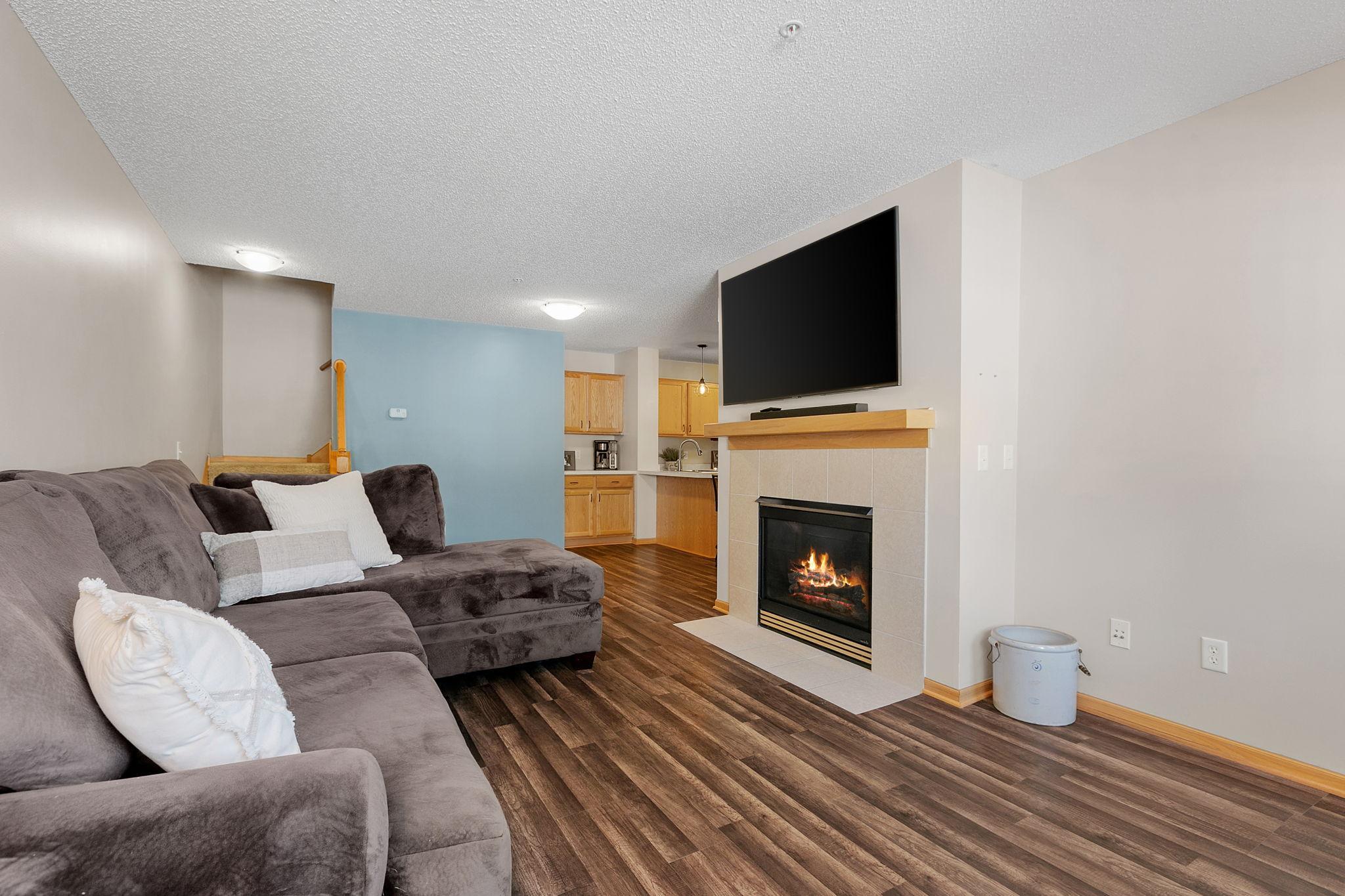 Living room with a gray sectional, lit fireplace, wall-mounted TV, and open kitchen.