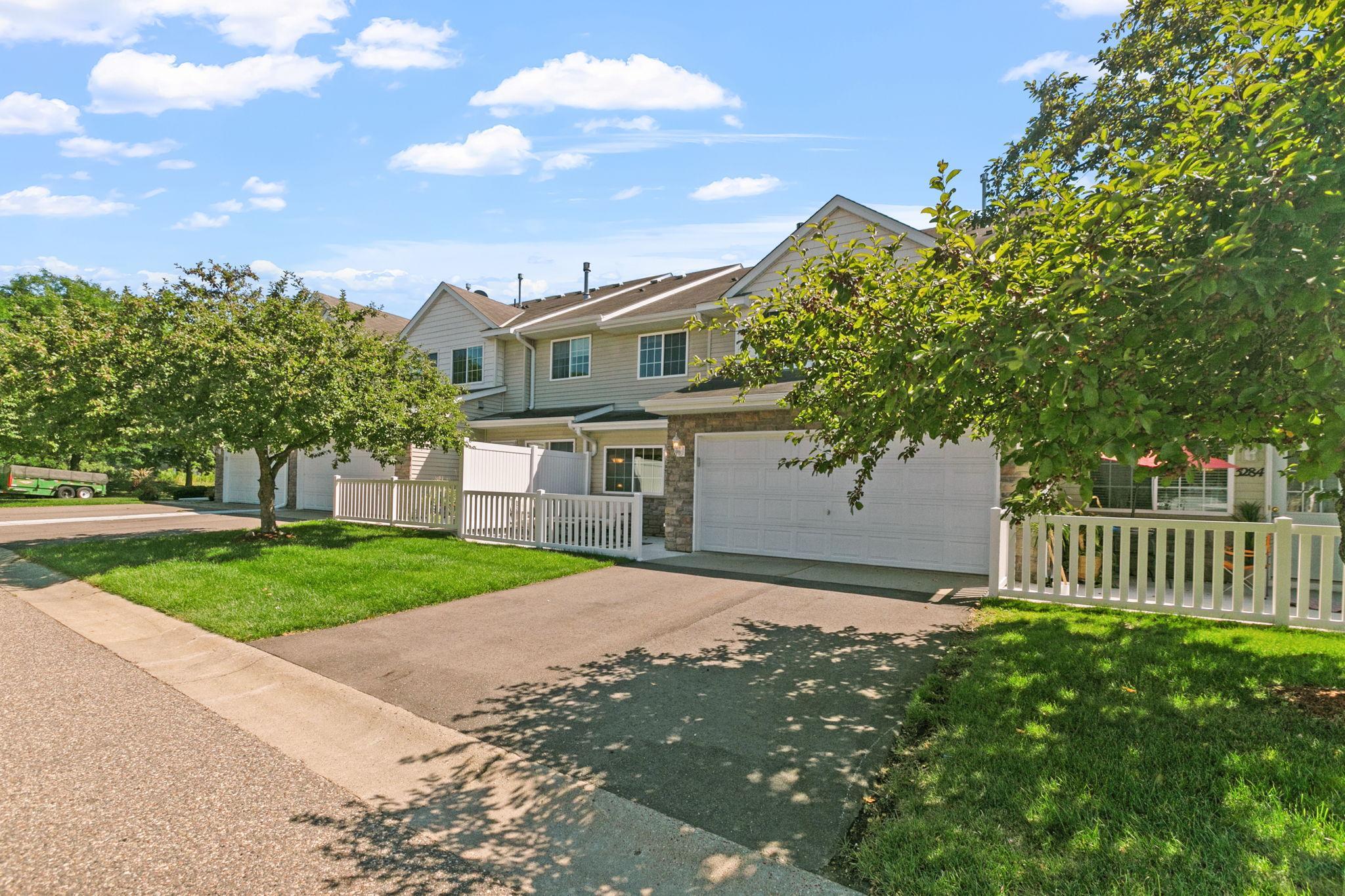 Attached townhomes with white picket fences, garages, and leafy trees on a sunny day.