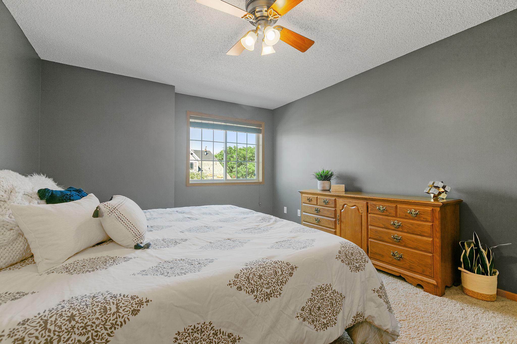 Furnished bedroom with gray walls, patterned white bedding, a wooden dresser, and a ceiling fan.