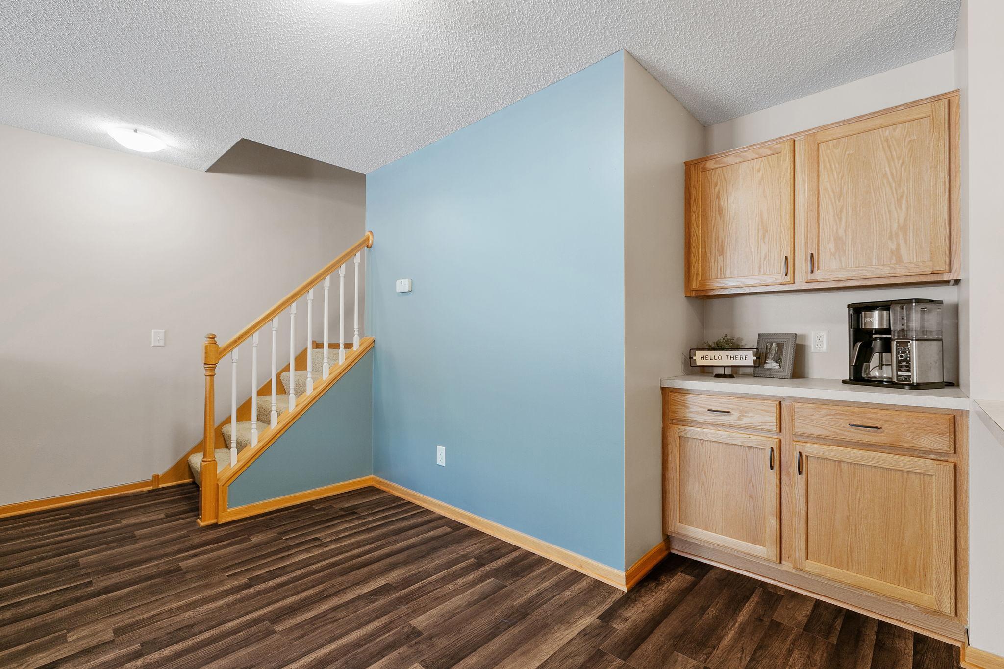 Built-in kitchenette with light wood cabinets and coffee maker beside a staircase and blue accent wall.