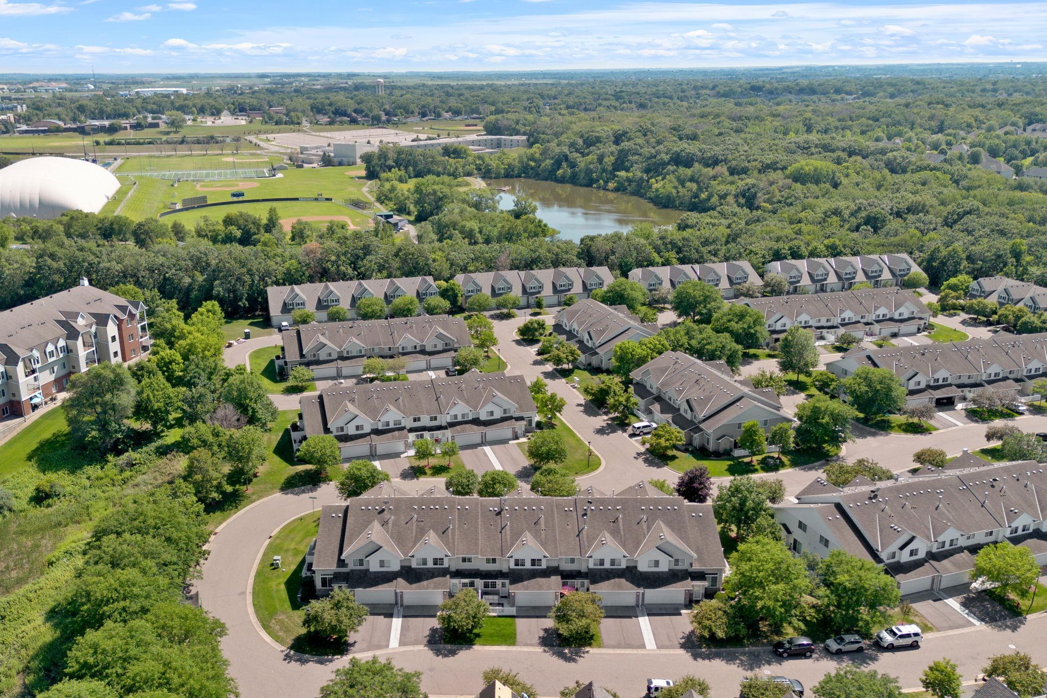 Aerial view of a townhome community beside a wooded pond and athletic fields.