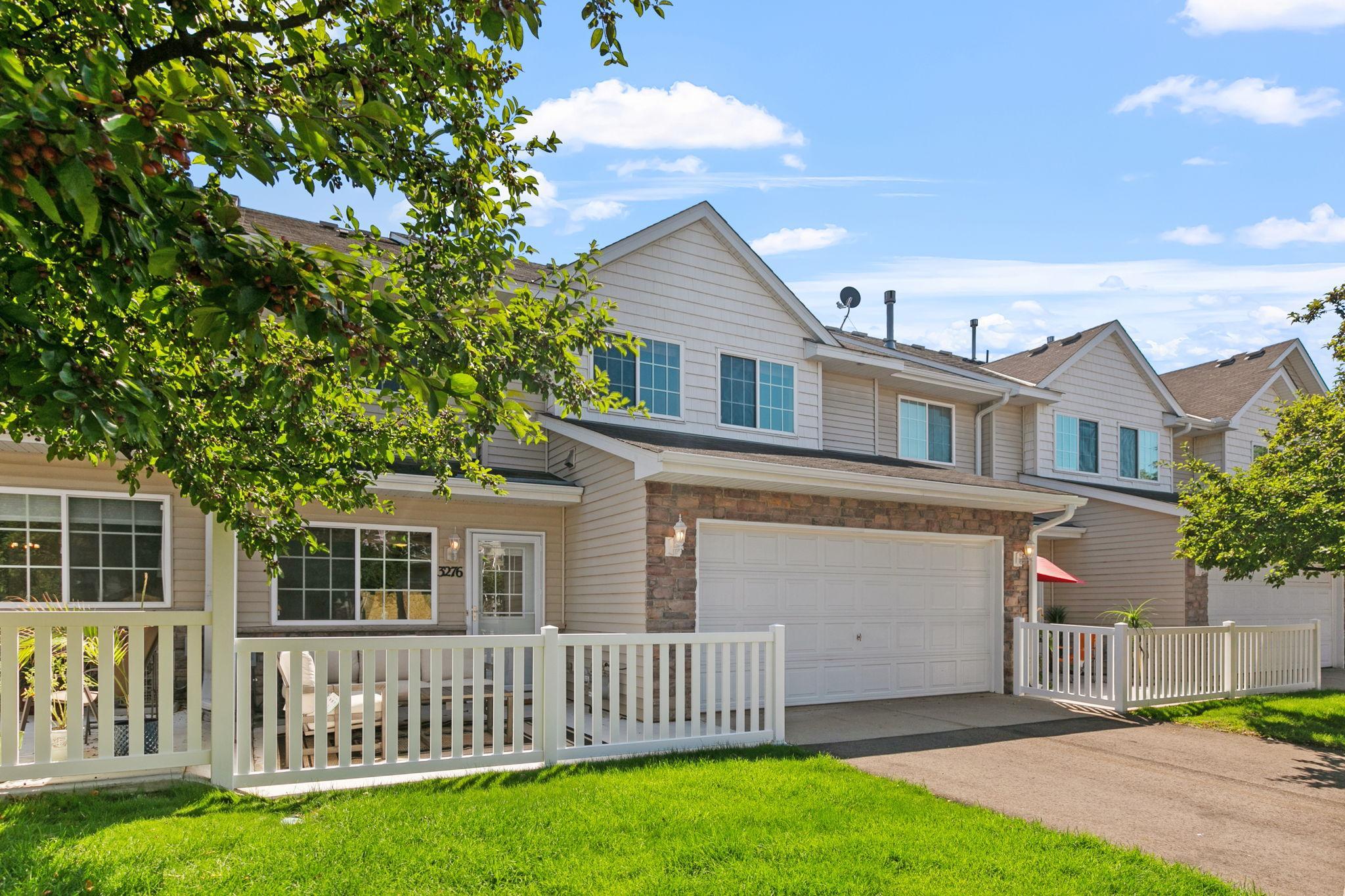 Attached townhouse with stone-faced garage, white fencing, and leafy tree in front.