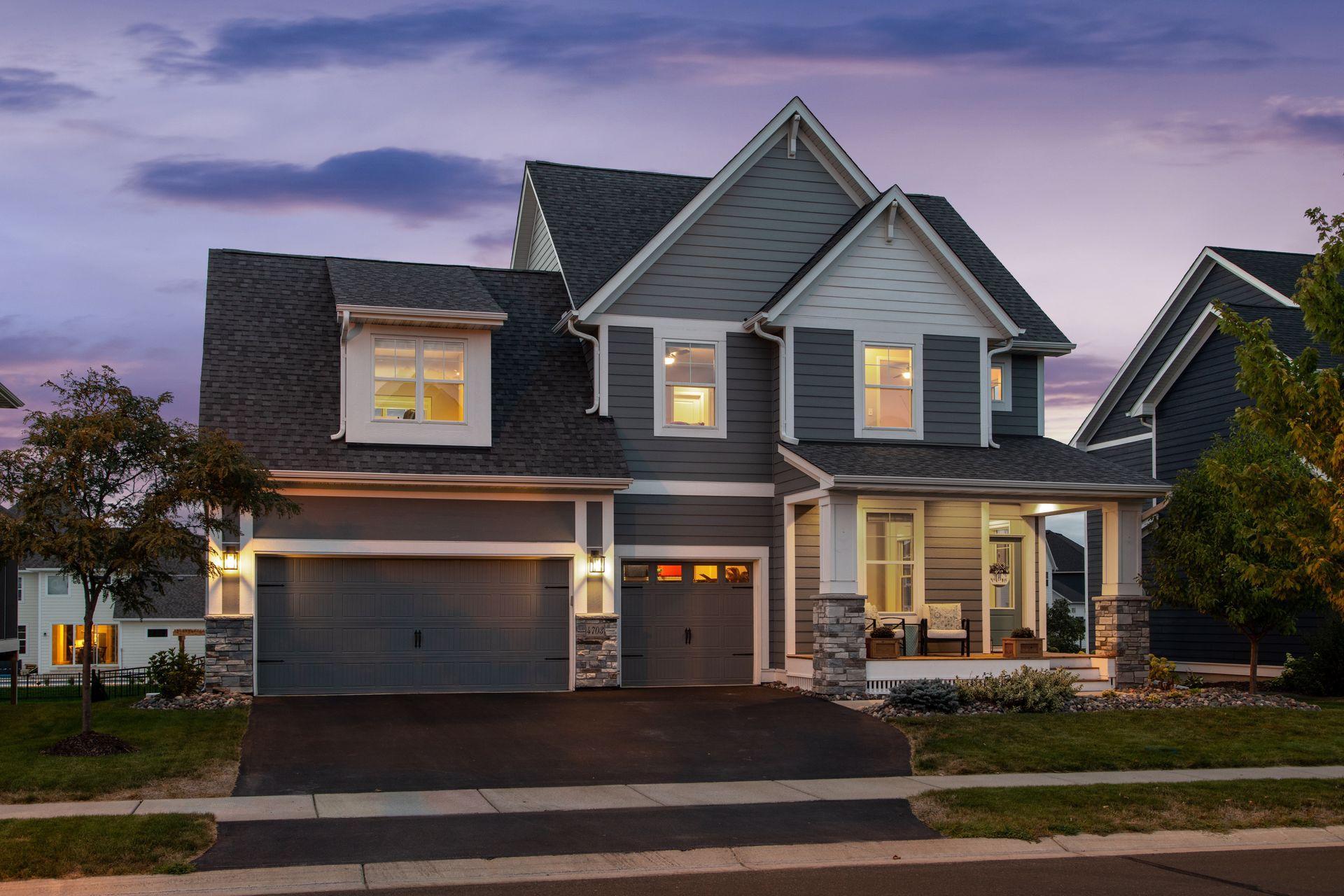 Gray two-story home with multiple gables, a three-car garage, and an illuminated covered porch at dusk.