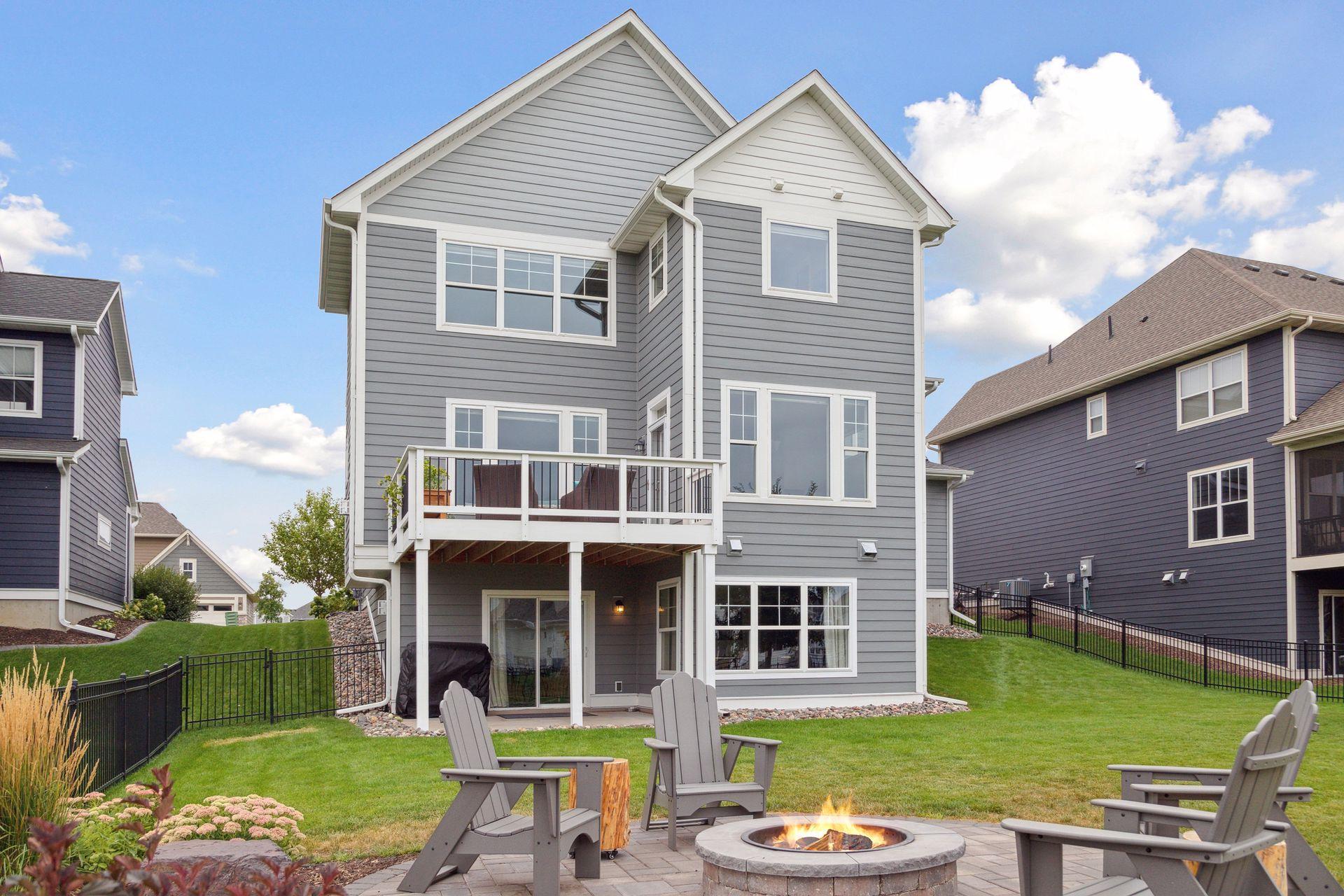 Gray two-story home behind a landscaped patio with Adirondack chairs and a circular fire pit.