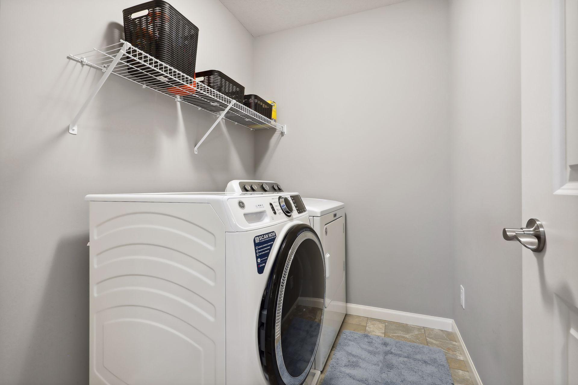 Laundry room with front-loading washer and dryer, wire shelf with baskets, and tiled floor.