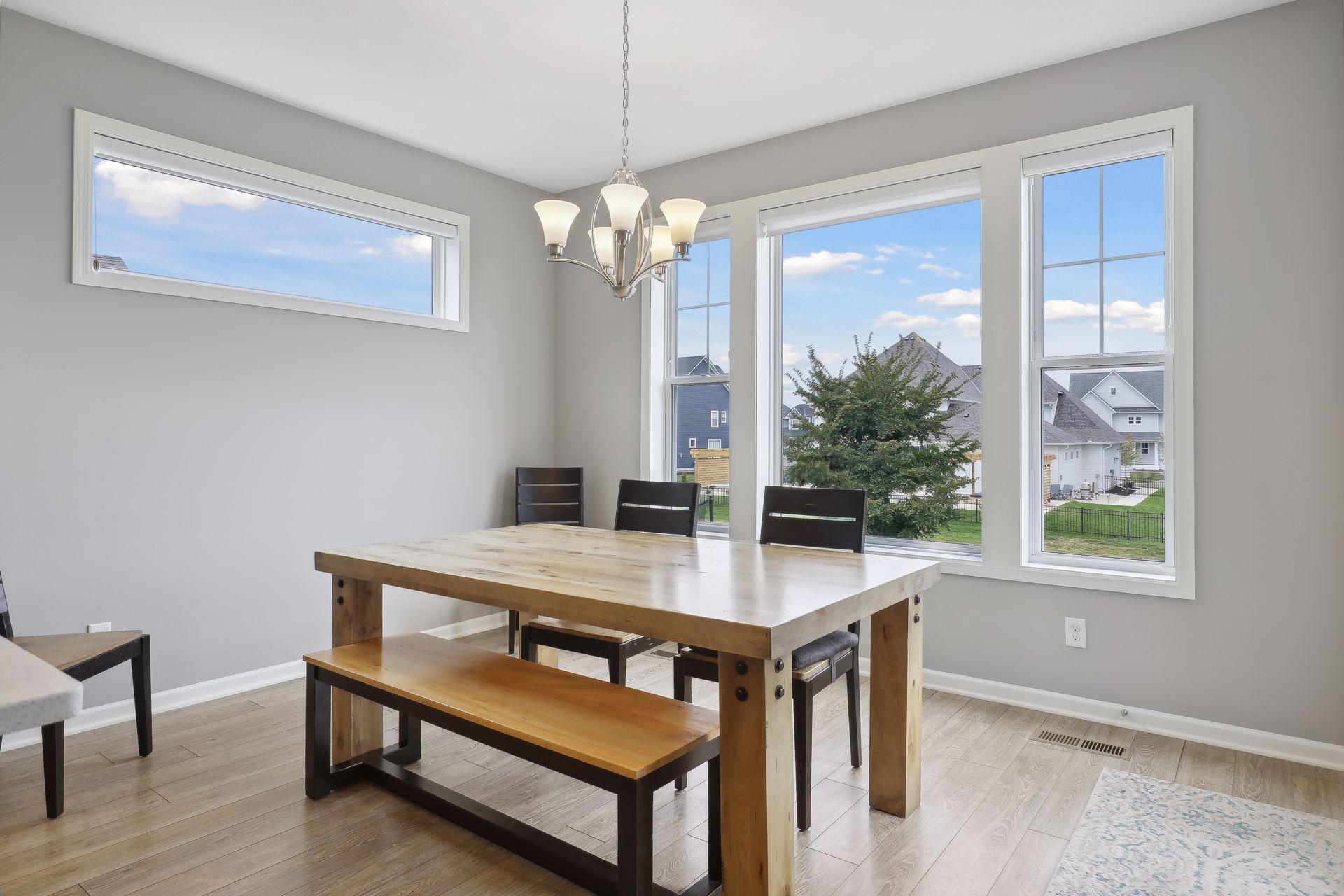 Dining room with a wood table and benches beneath a chandelier, beside large windows overlooking neighboring homes.
