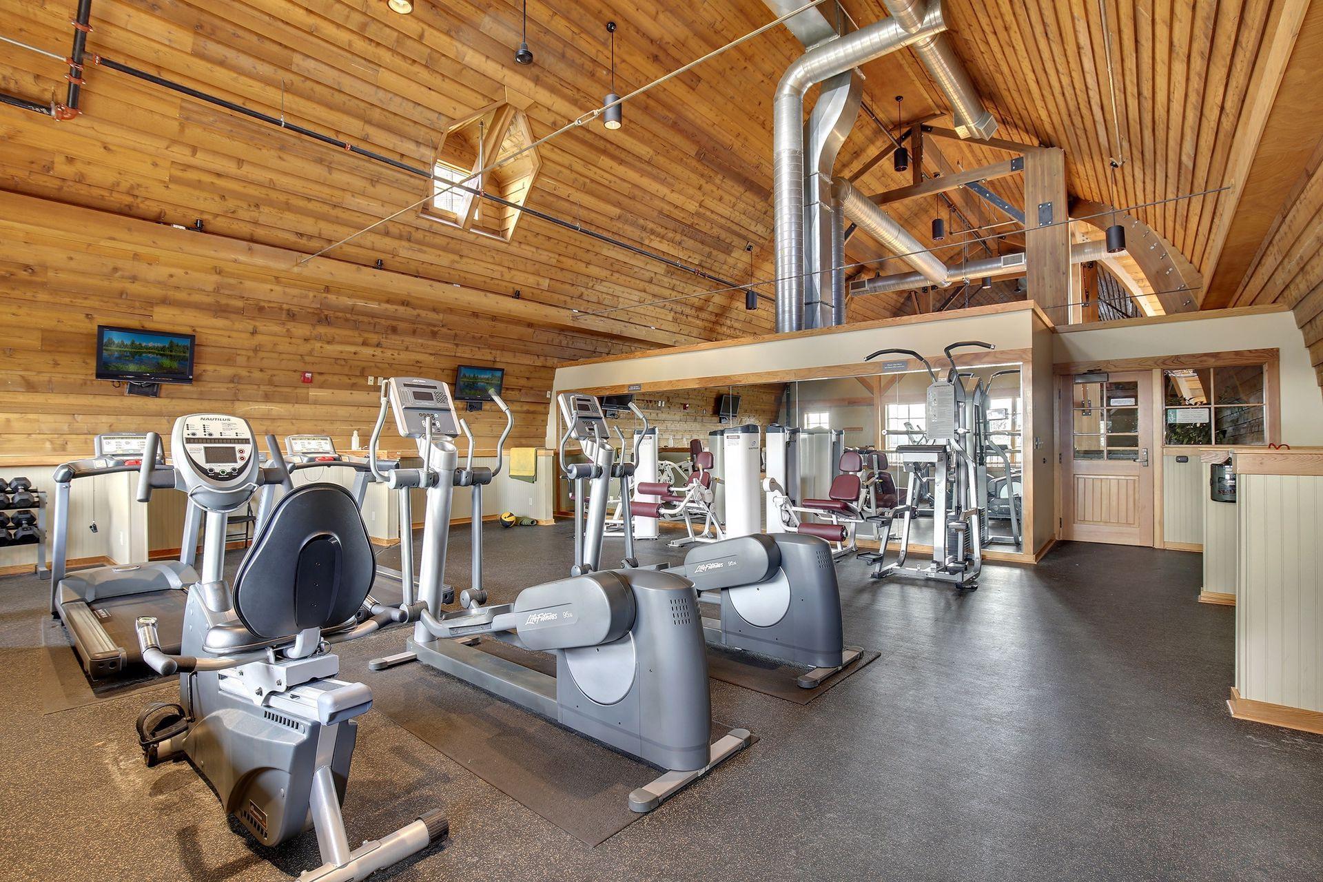 Gym with cardio and weight equipment, mirrored wall, and vaulted wood-plank ceiling.