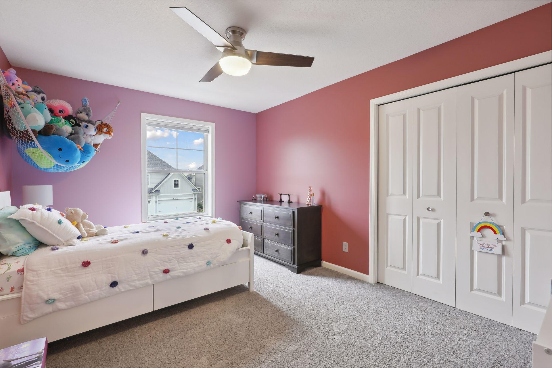 Bedroom with lavender and coral walls, a white pom-pom bed, stuffed animals, and white closet doors.