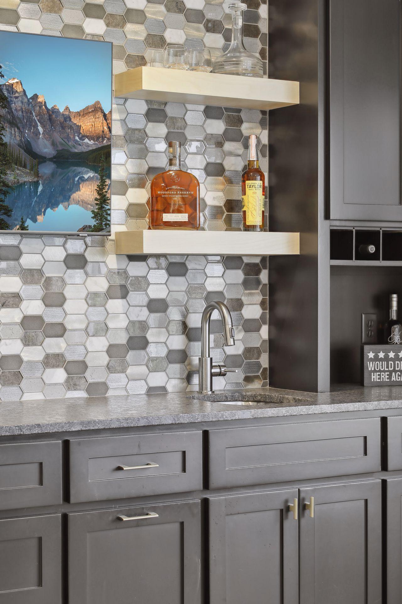 Kitchen bar with hexagonal tile backsplash, floating shelves, gray cabinets, and a tall sink faucet.