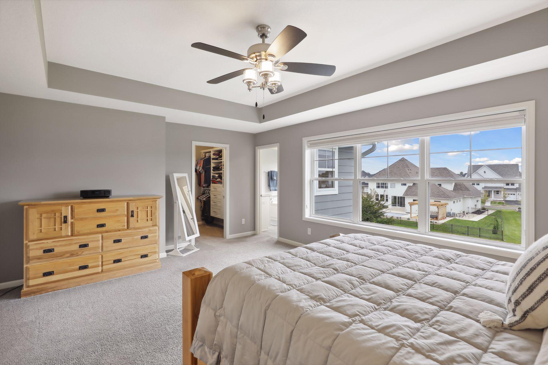 Bedroom with a tray ceiling, ceiling fan, and large window overlooking neighboring homes.