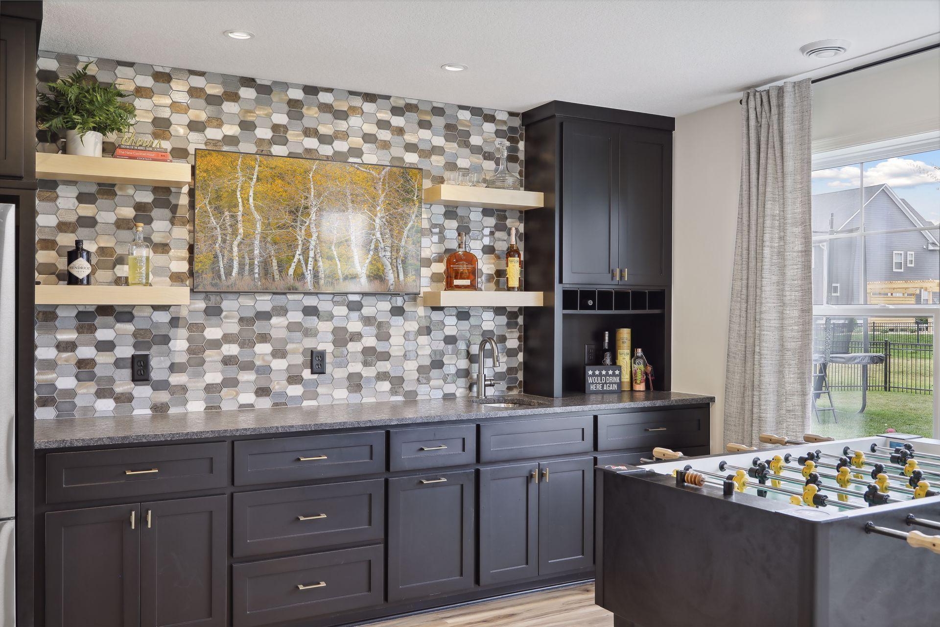 Basement wet bar with a hexagonal tile backsplash and foosball table beside a large window.