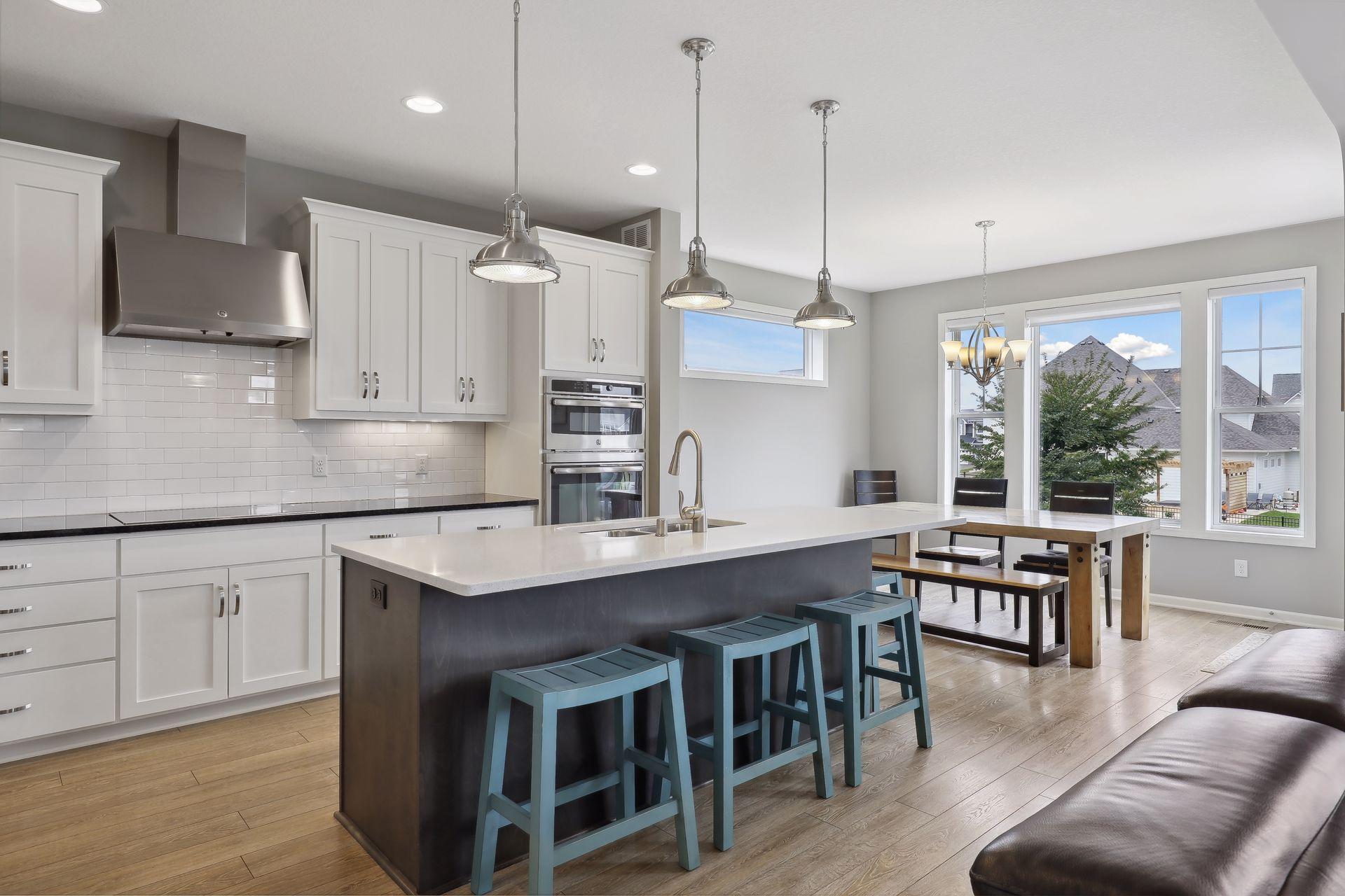 Open-plan kitchen with a dark island, teal stools, and a dining area beside large windows.