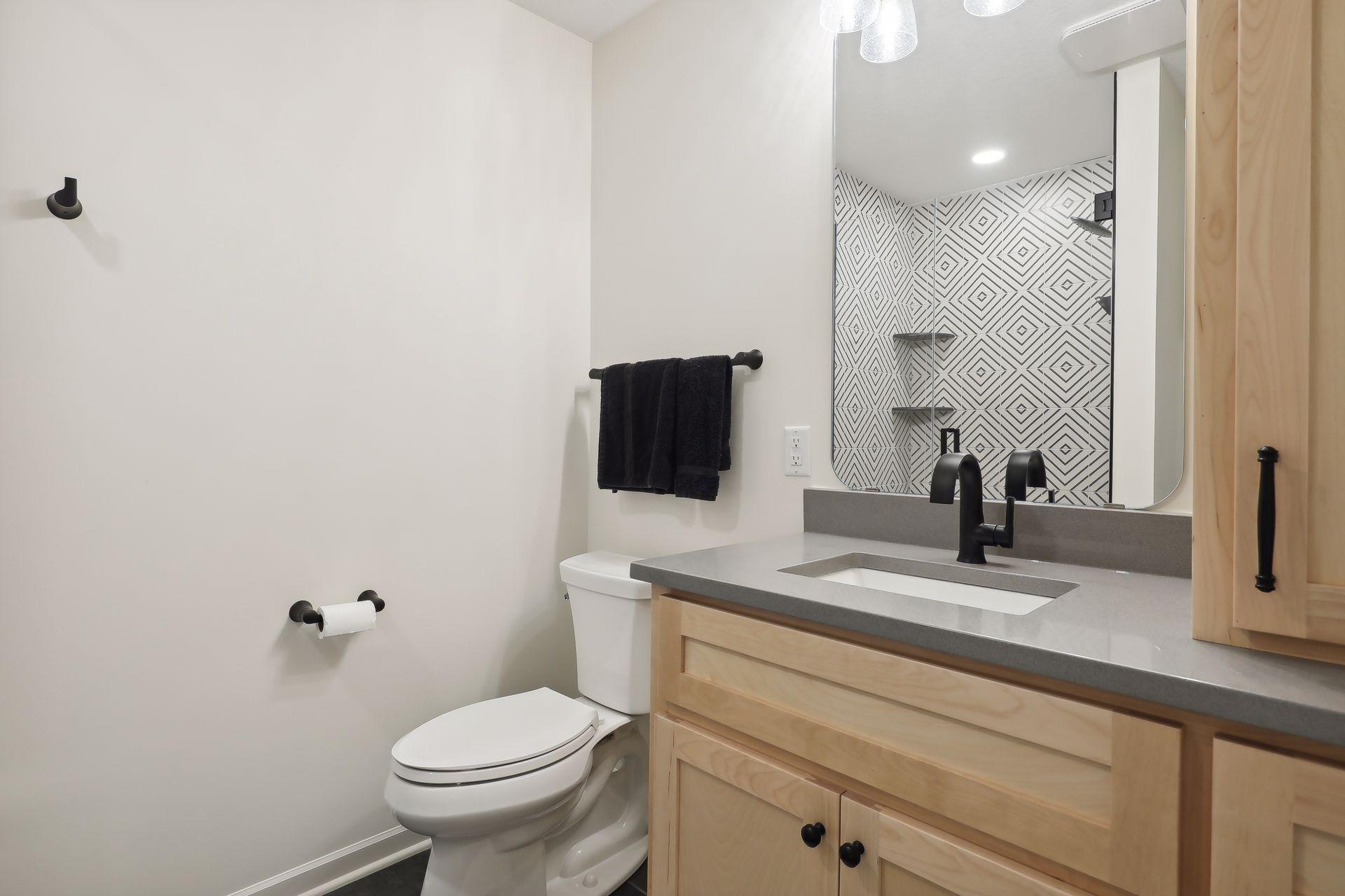 Bathroom with light wood vanity, black faucet, and geometric-tile glass shower.