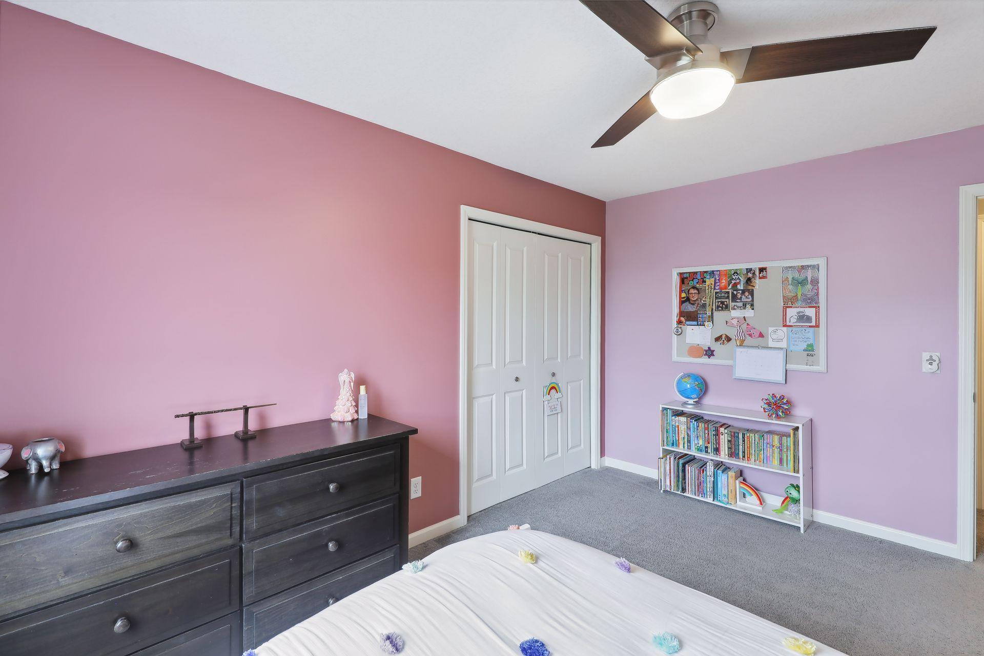 Bedroom with pink and lavender walls, a dark dresser, white closet doors, and a bookshelf filled with books.