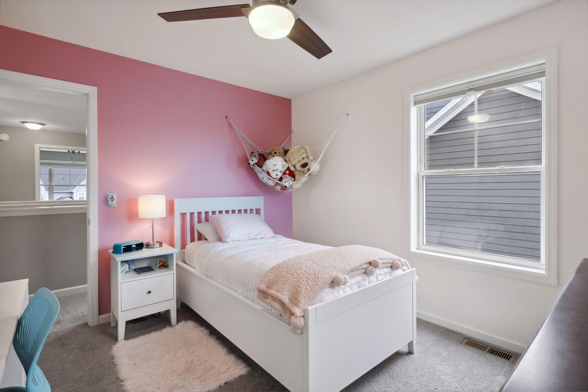 Bedroom with a white bed, pink accent wall, hanging plush toys, and a large window.