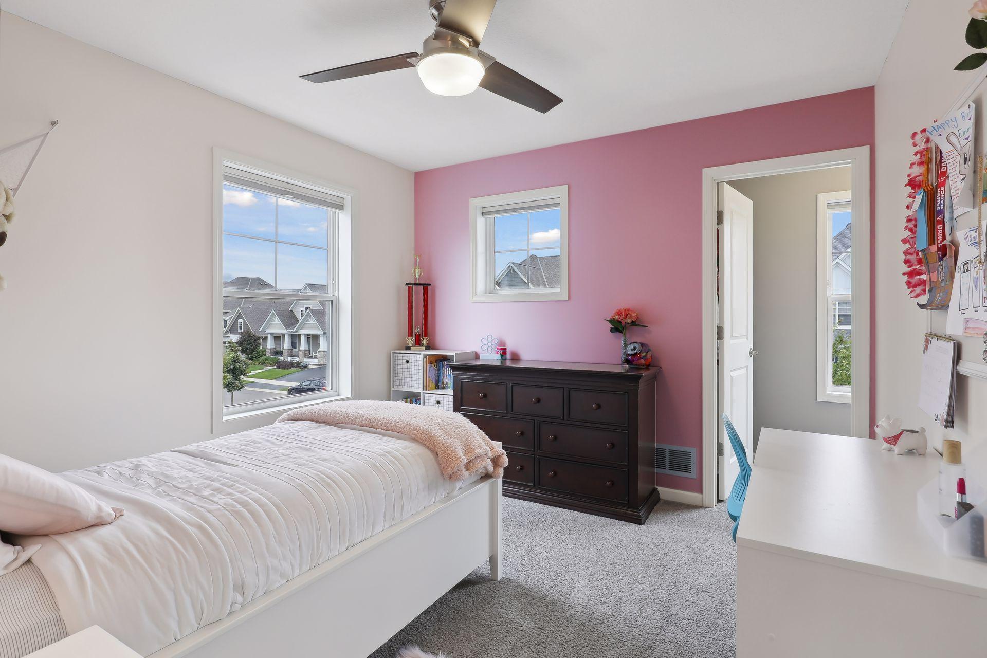 Bedroom with a pink accent wall, white bed, dark dresser, and windows overlooking neighboring homes.