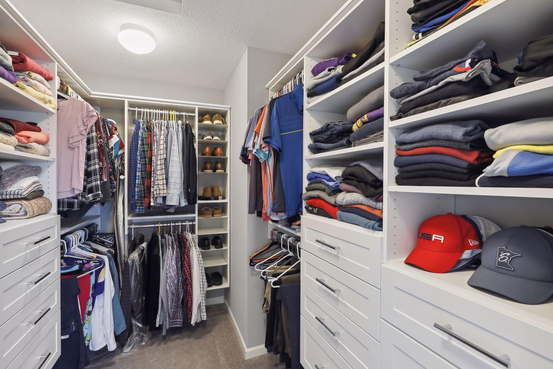 Walk-in closet with white built-in drawers, shelves of folded clothes, hanging garments, and shoe cubbies.