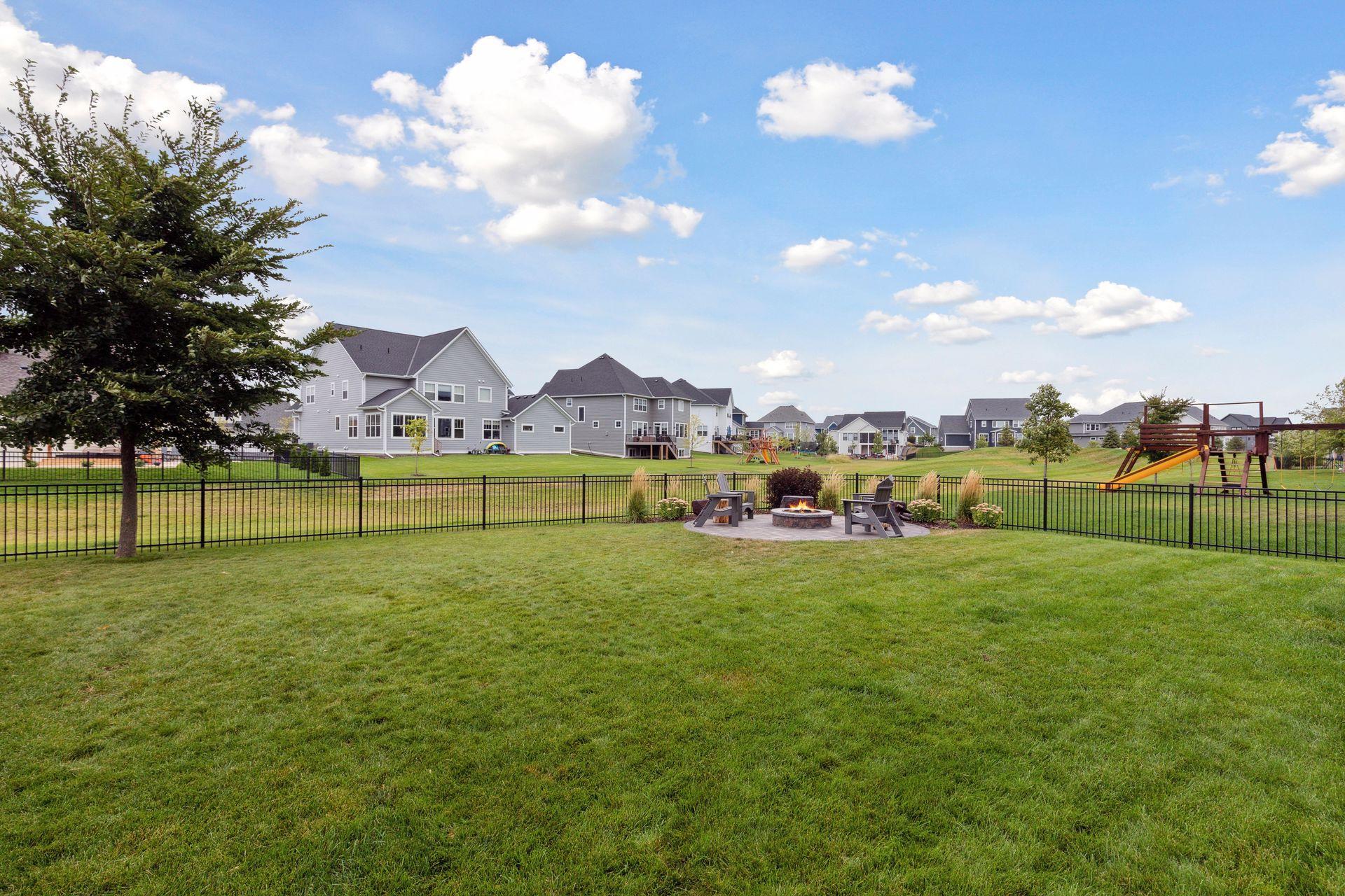 Fenced backyard with a fire pit seating area, neighboring homes, and a playground beyond the lawn.