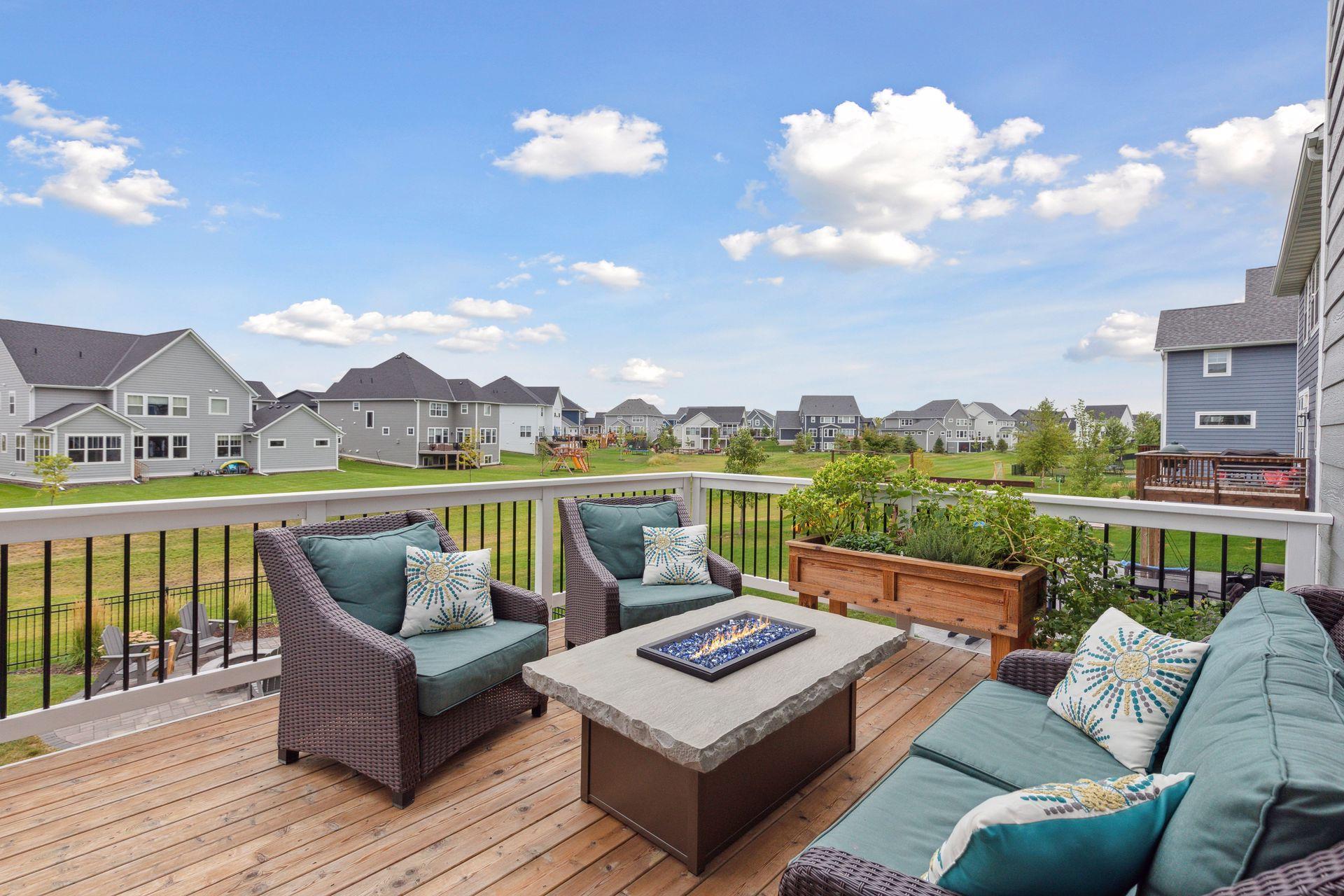 Furnished wooden deck with teal-cushioned seating and a fire table overlooking a neighborhood of homes.