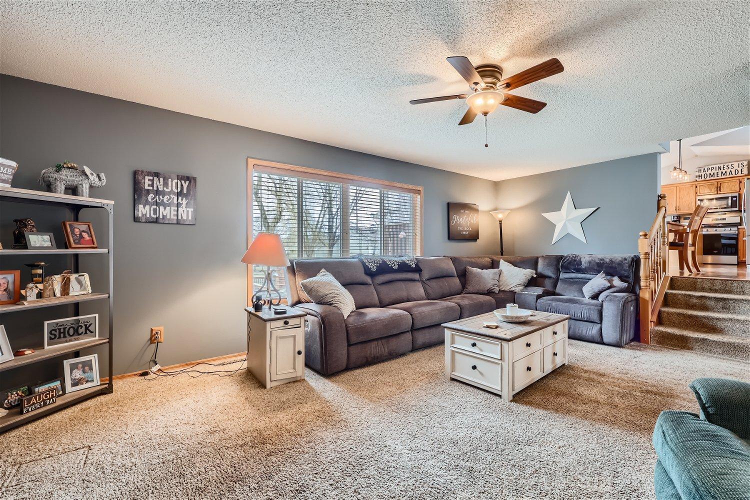 Living room with a gray sectional, cream coffee table, carpeted floor, and ceiling fan.