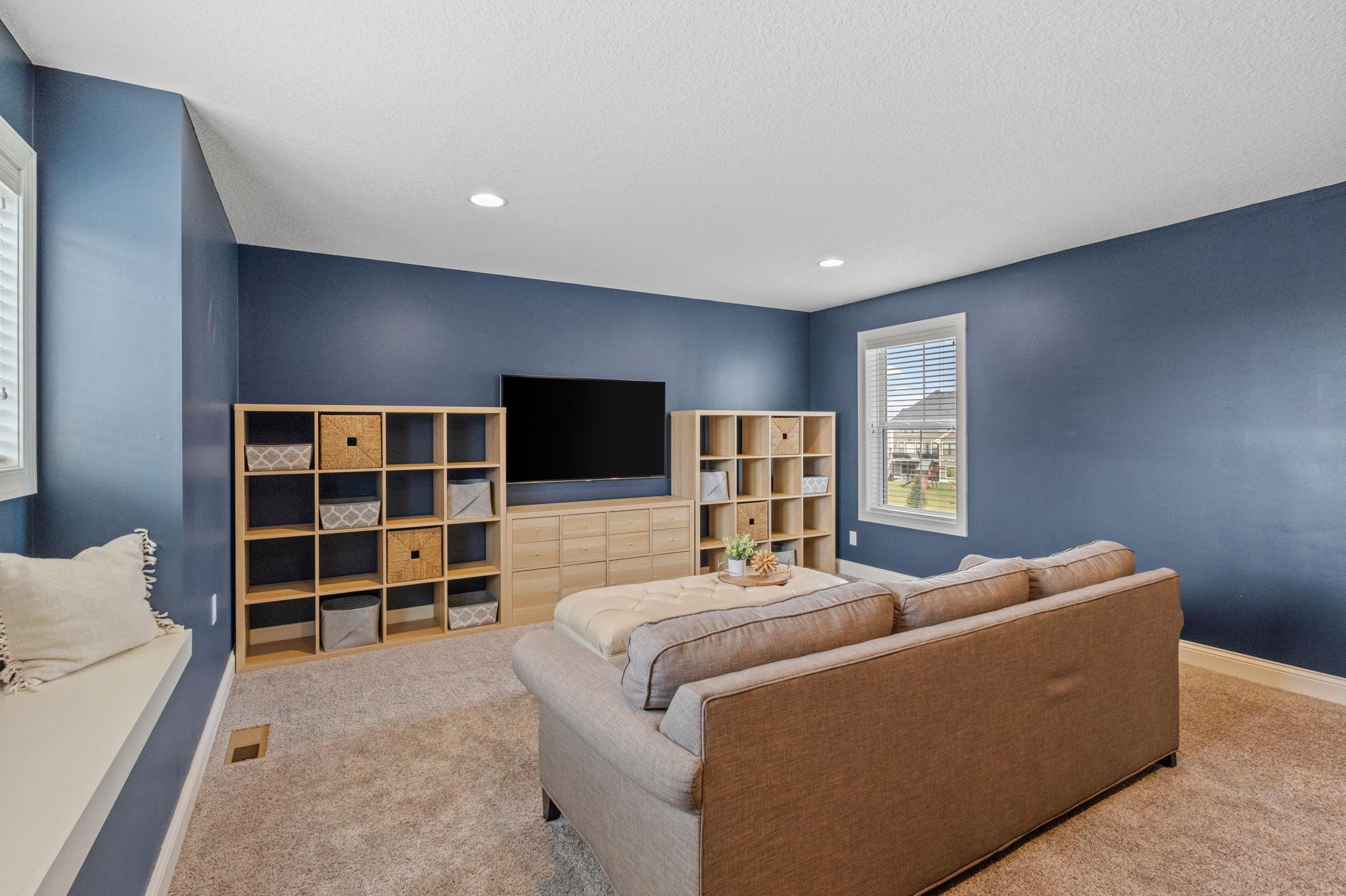 Furnished family room with blue walls, a large sectional sofa, wall-mounted TV, and light wood shelving.