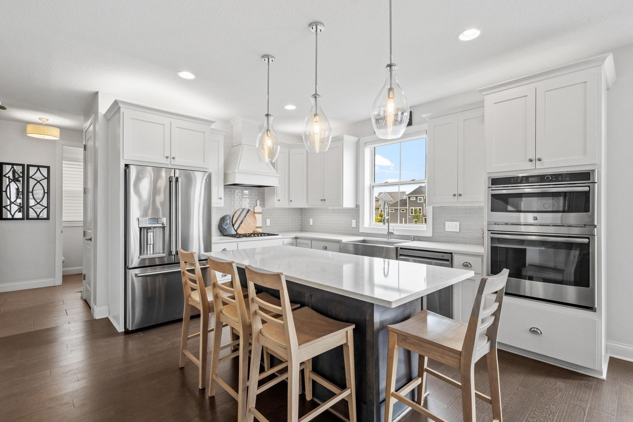 Bright white kitchen with a large island, wooden stools, stainless appliances, and glass pendant lights.
