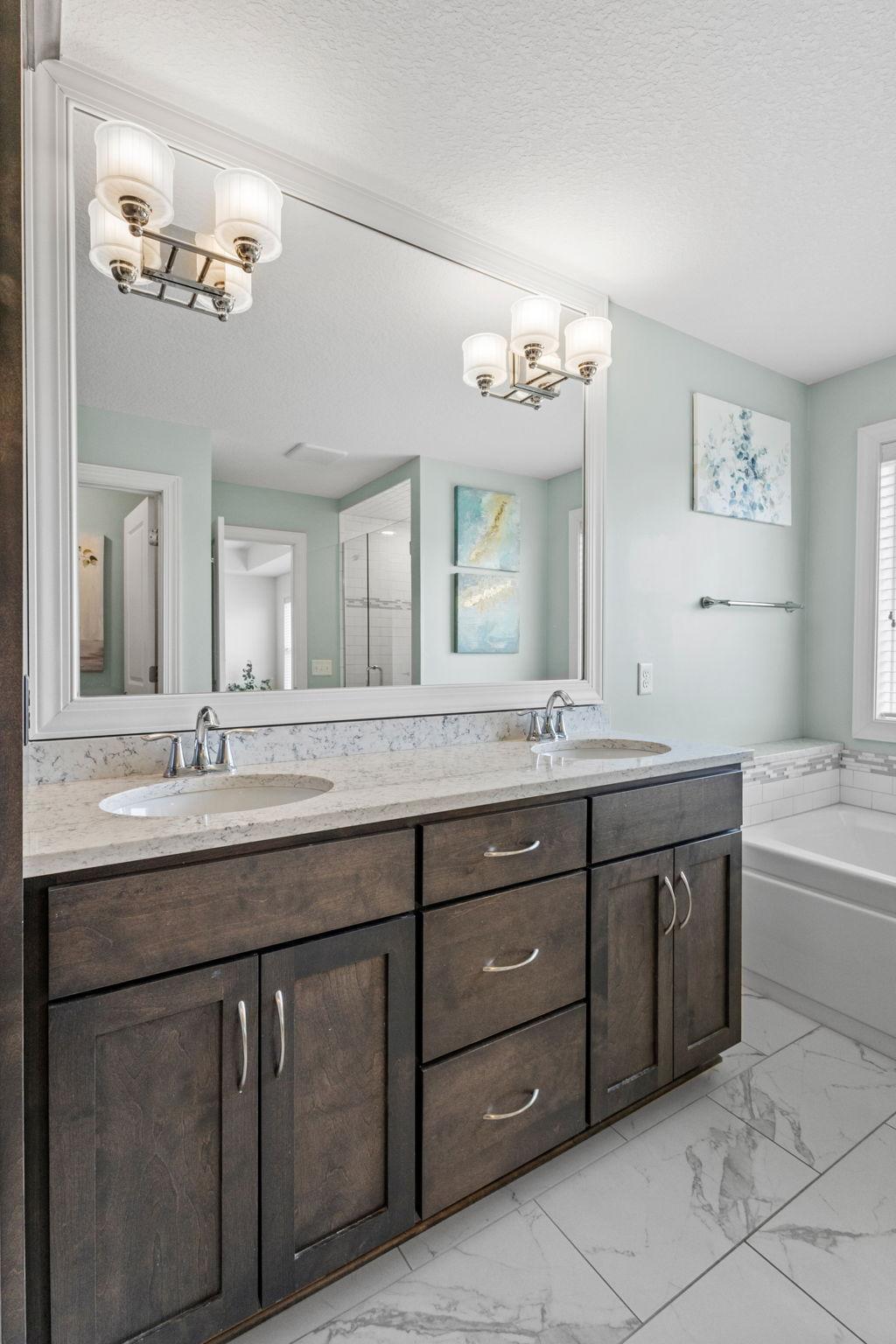 Bathroom with dark double vanity, large mirror, marble-look counter, and bathtub with tiled surround.