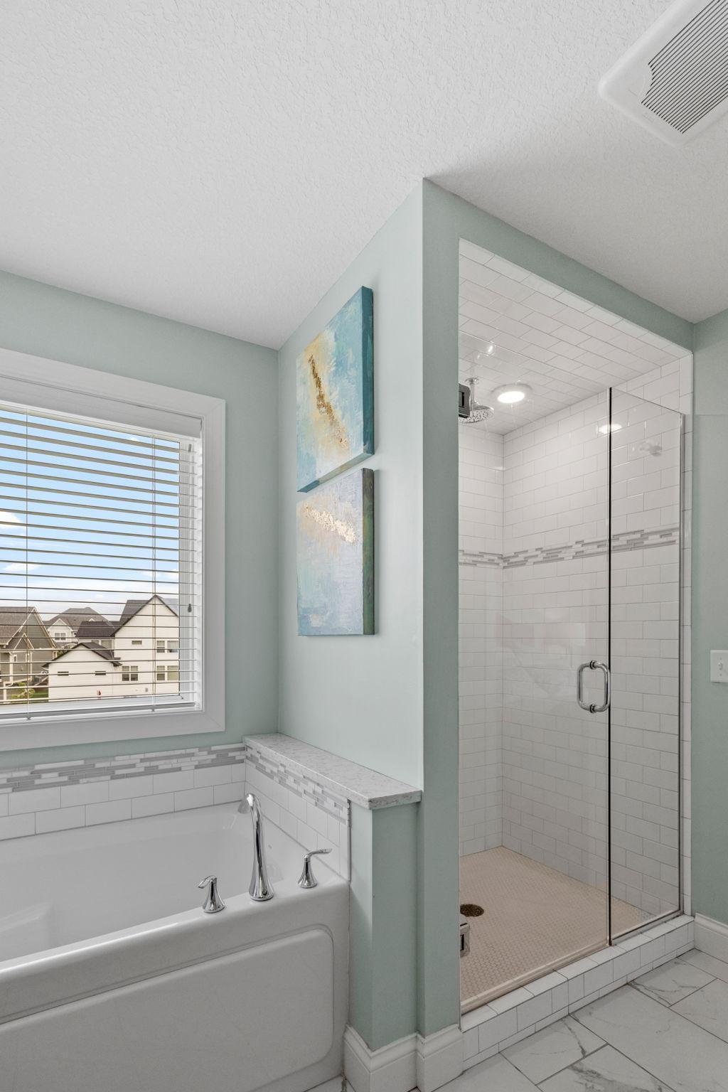 Bathroom with a soaking tub beside a glass-enclosed tiled shower and a window overlooking nearby homes.