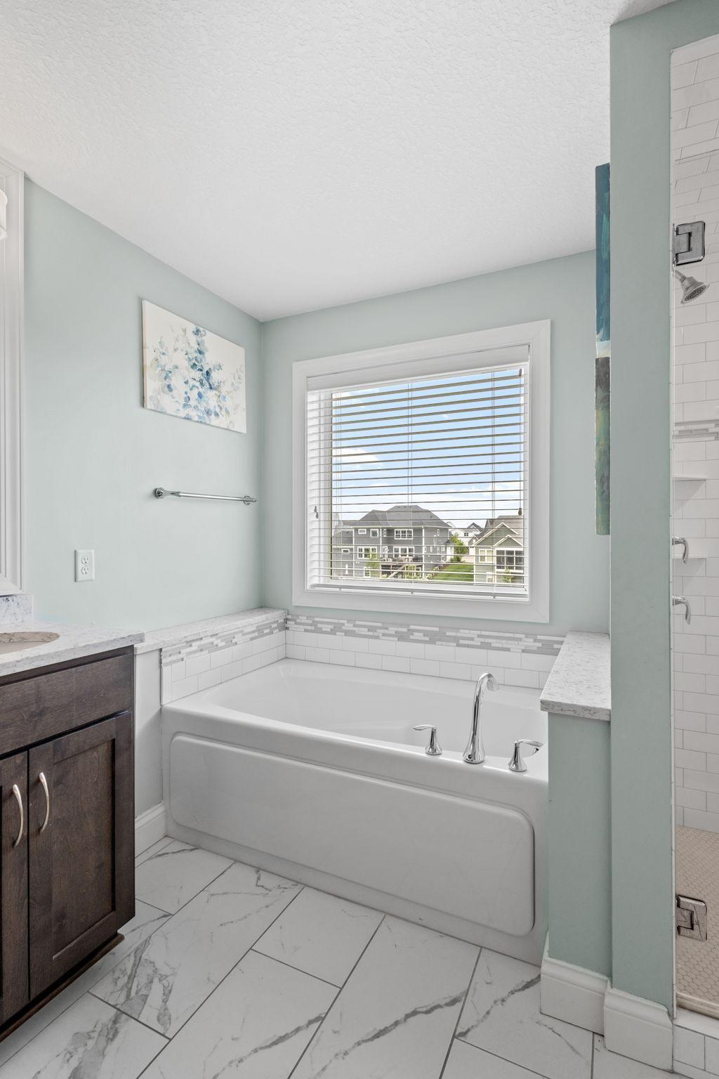 Bathroom with a soaking tub beneath a window, marble-look tile, and a dark wood vanity.