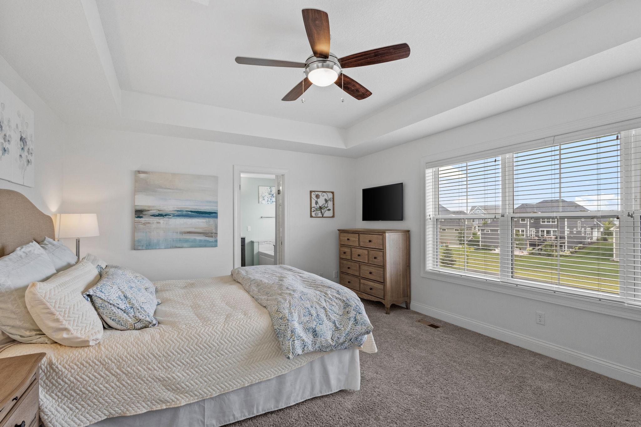 Furnished bedroom with a tray ceiling, ceiling fan, and large windows overlooking neighboring homes.