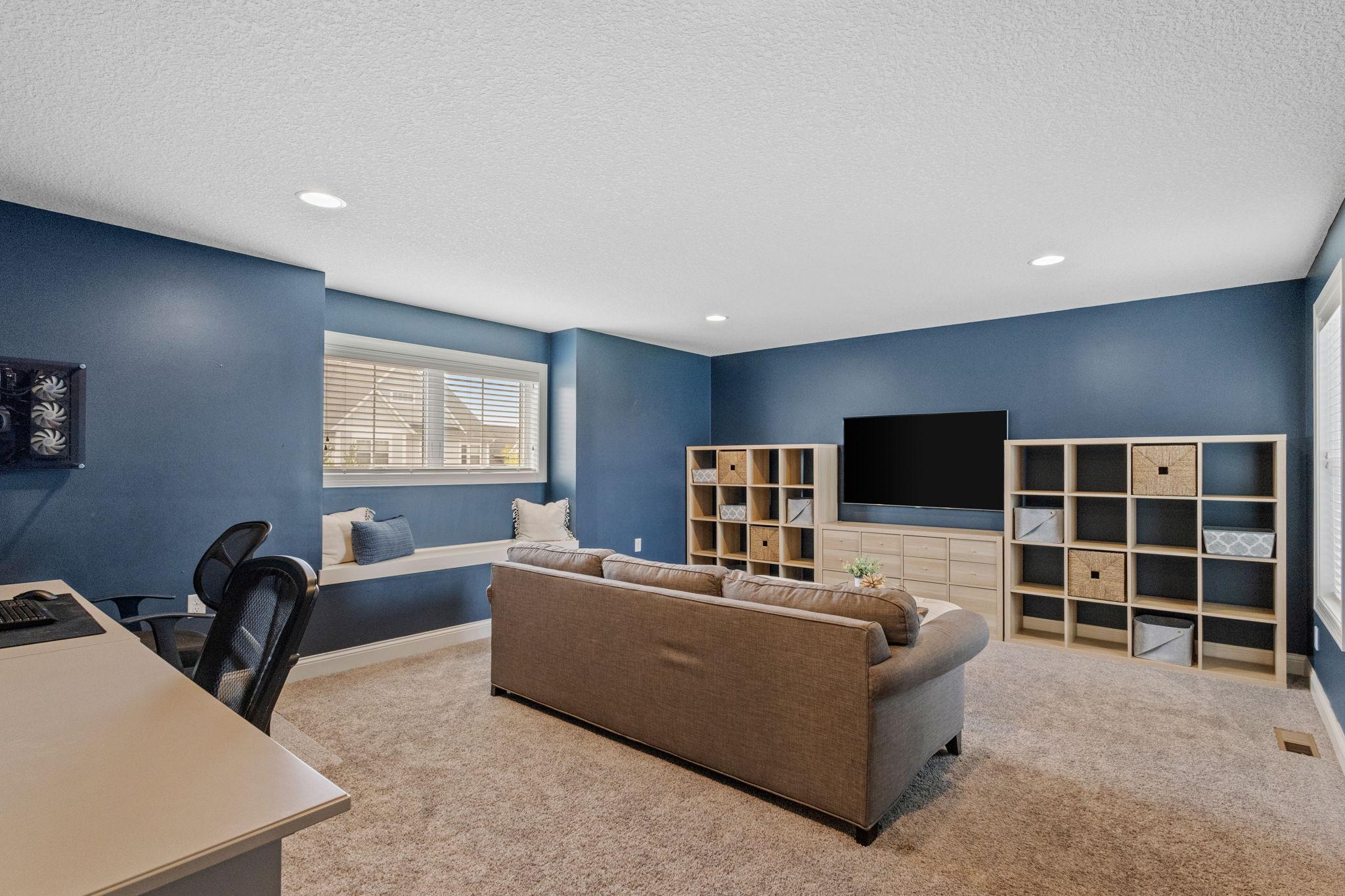 Basement family room with blue walls, brown sectional, large TV, built-in window seat, and desk area.