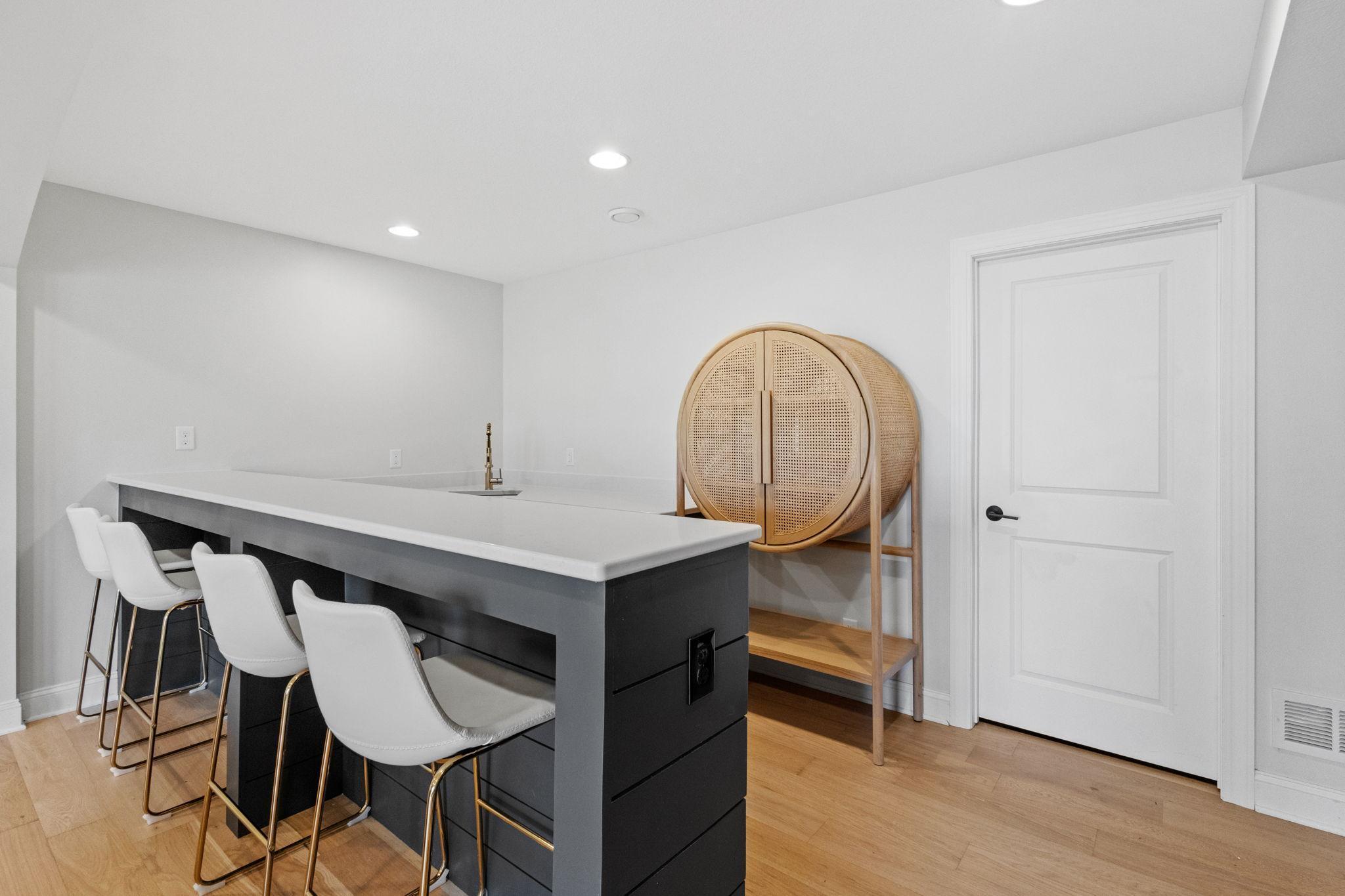 Basement bar with a dark island, three white stools, brass faucet, and round rattan cabinet.