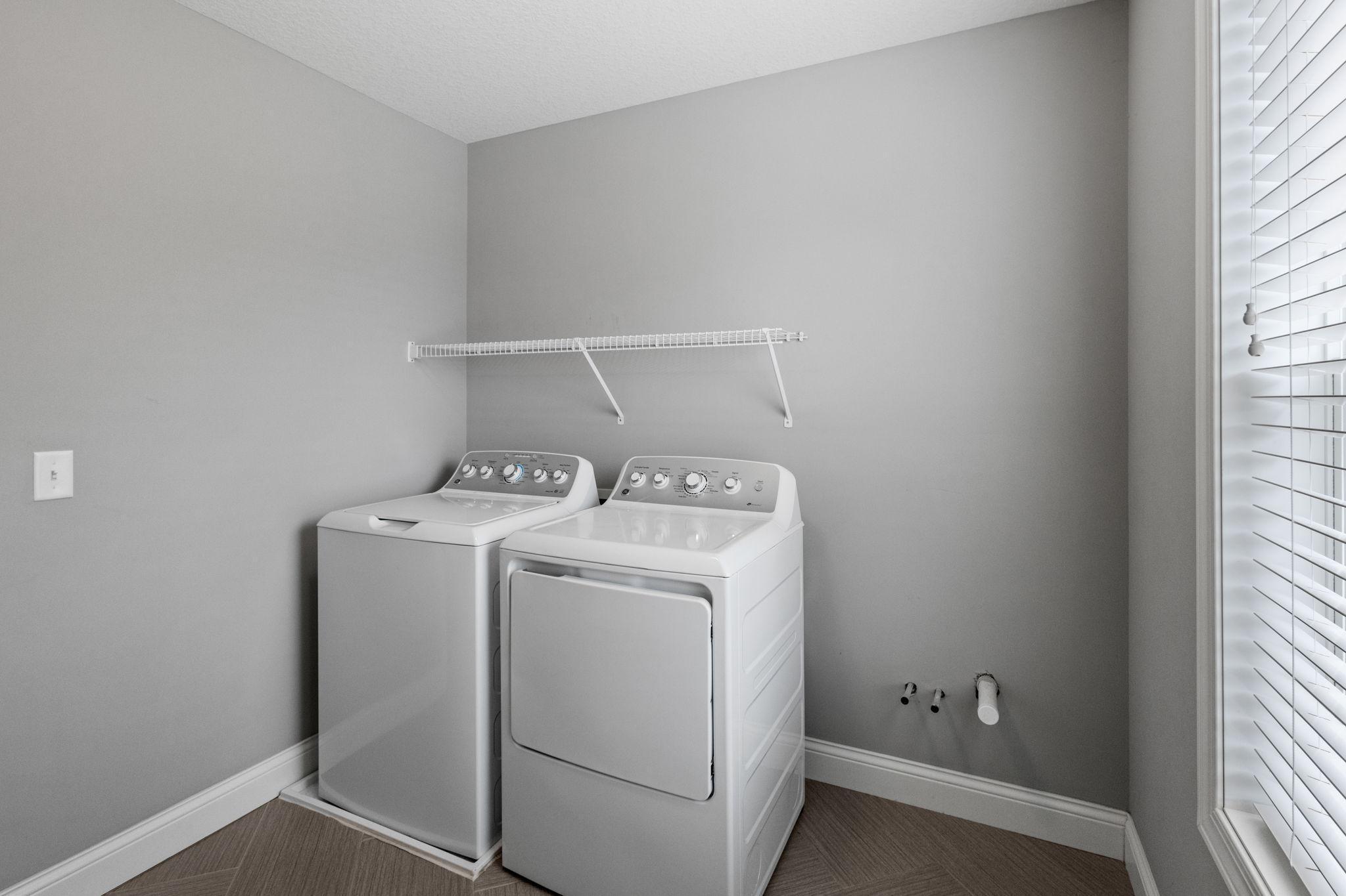Laundry room with white washer and dryer, wire shelf, and large window with blinds.