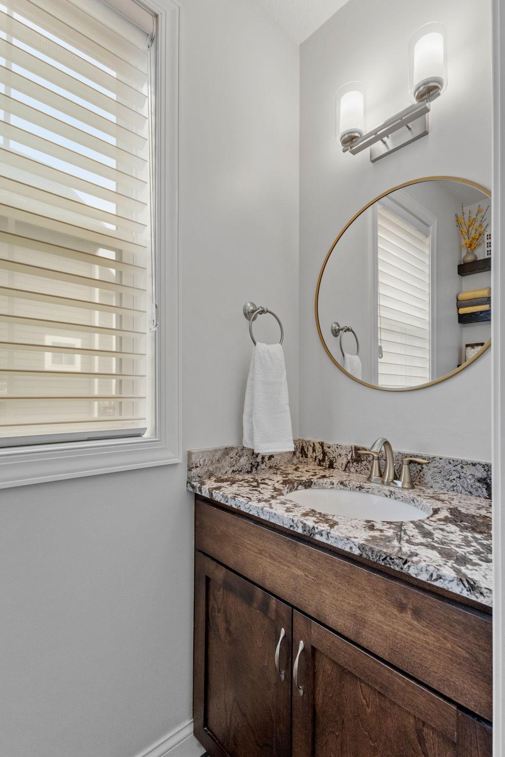 Bathroom vanity with a dark wood cabinet, stone countertop, round mirror, and window blinds.