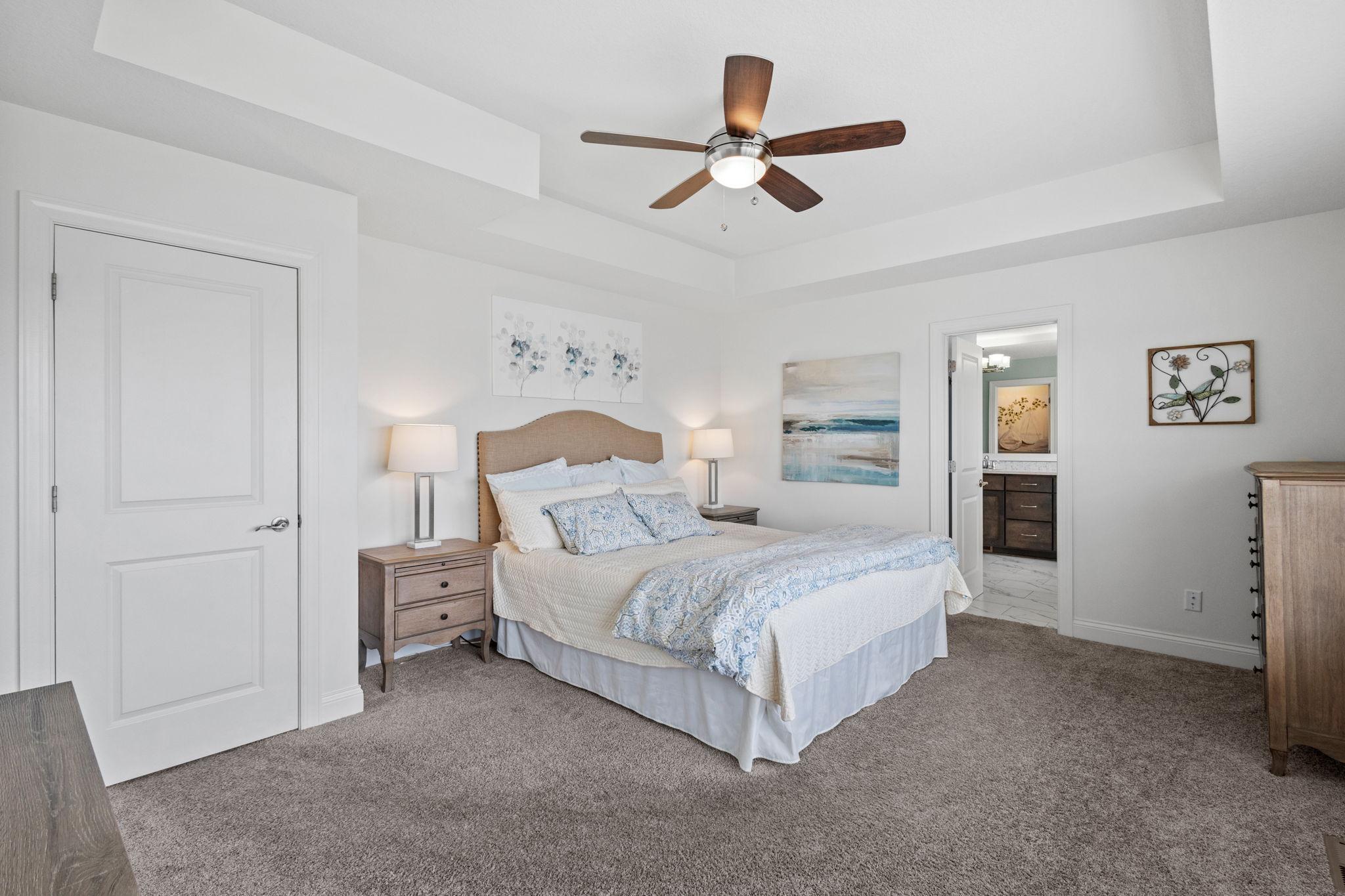 Furnished bedroom with a tray ceiling, wood-blade fan, and open doorway to the adjoining bathroom.