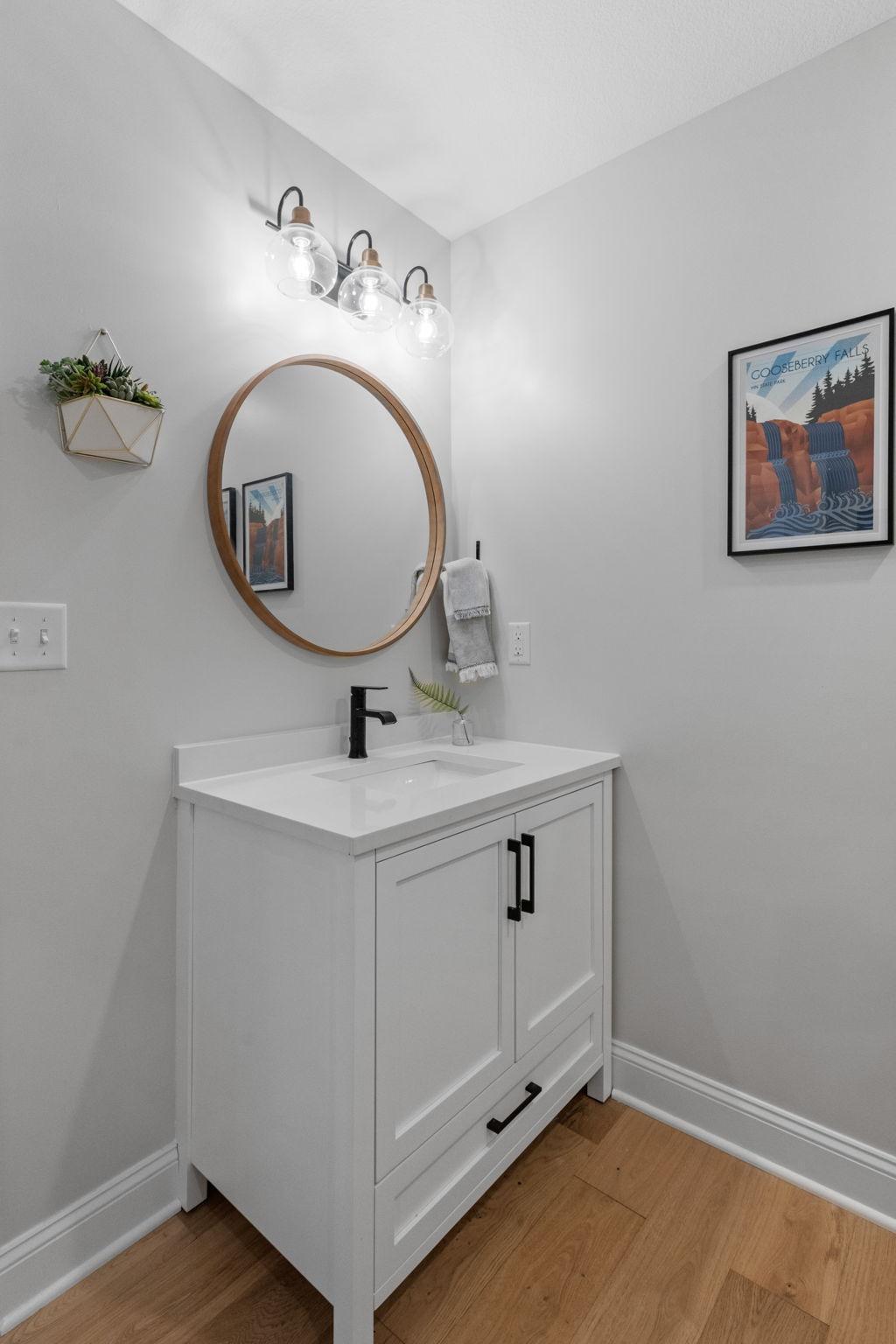 Bathroom vanity with a round wood-framed mirror, black faucet, and framed Gooseberry Falls print.