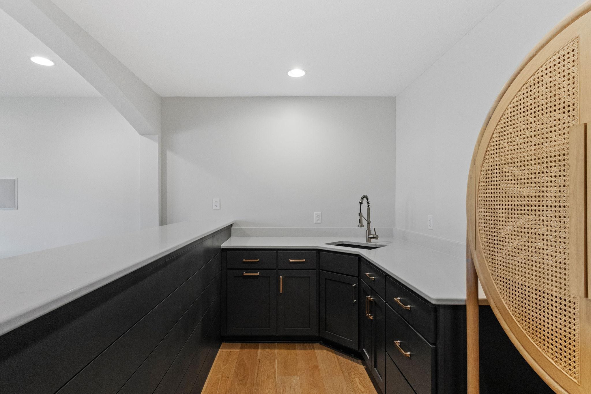 Modern kitchen with dark cabinets, white counters, a sink, and a cane-backed chair in the foreground.