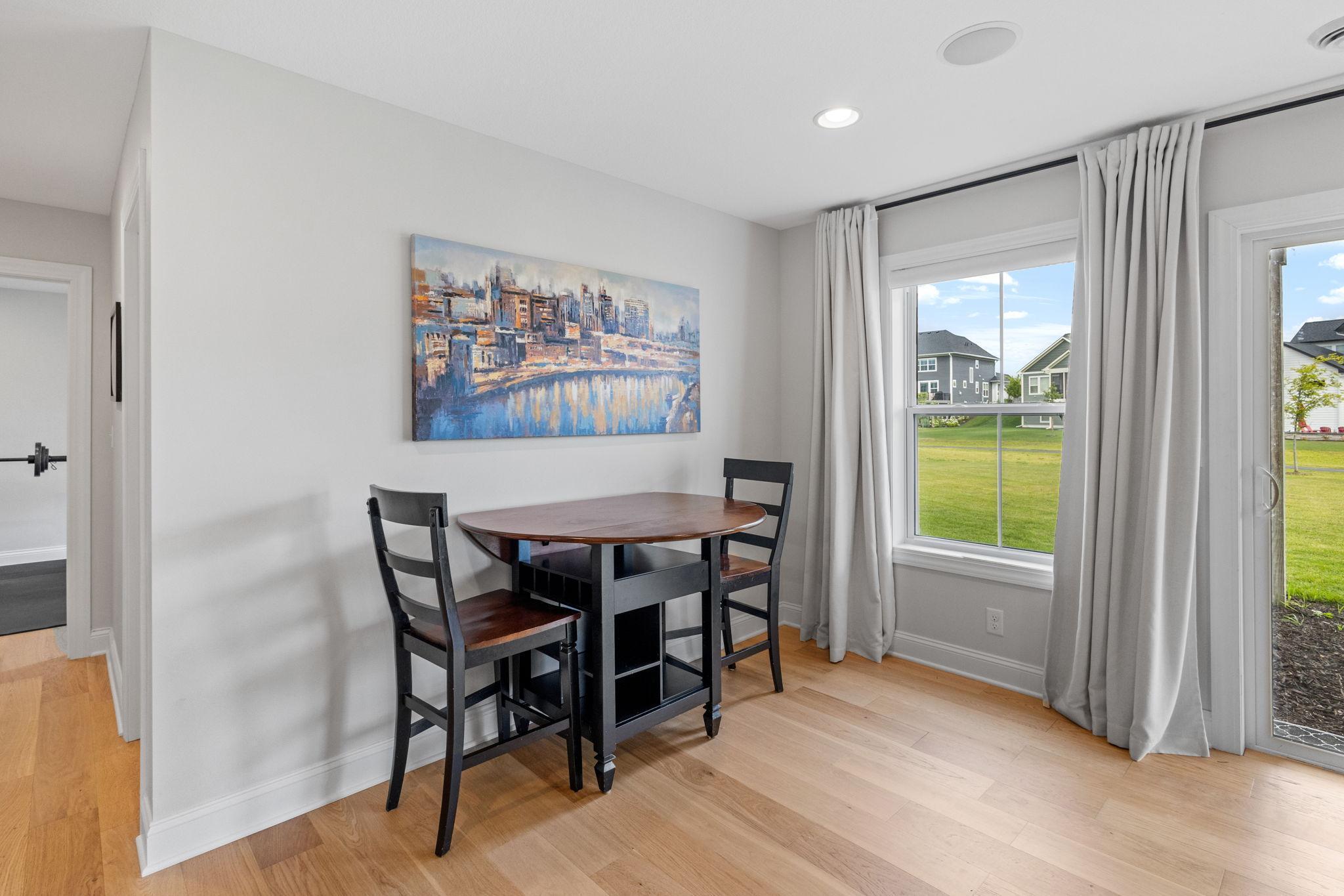 Dining nook with a round wooden table, two dark chairs, and curtained windows overlooking a lawn.