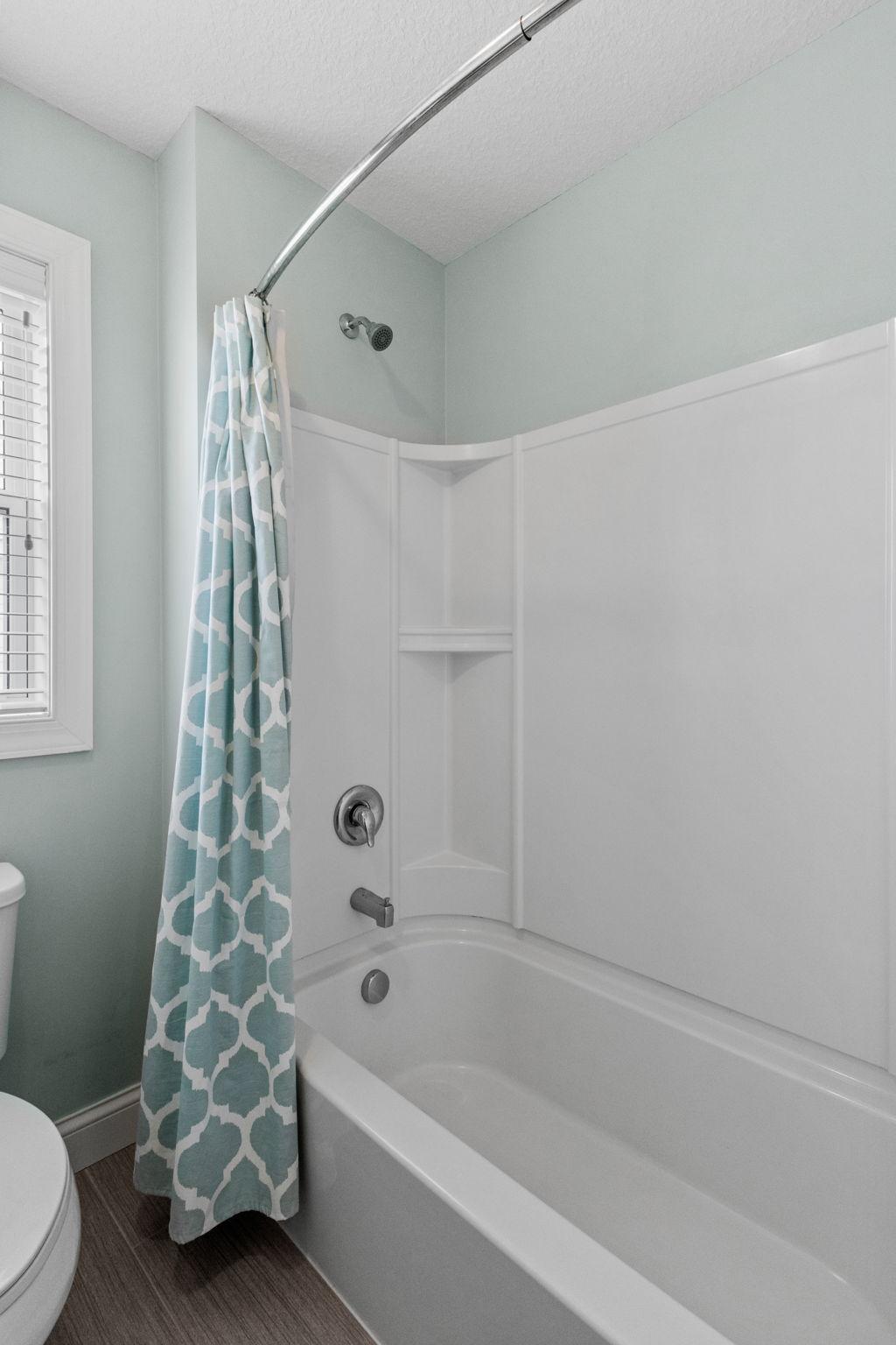 Bathroom with a white tub and shower, patterned curtain, pale aqua walls, and a window with blinds.