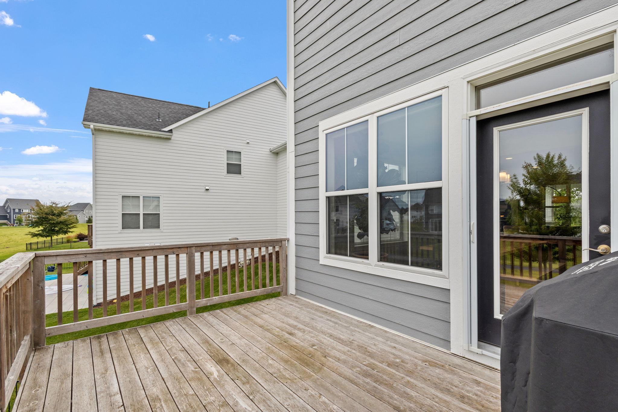 Wooden deck beside a gray-sided house, with a glass door and neighboring homes beyond the railing.