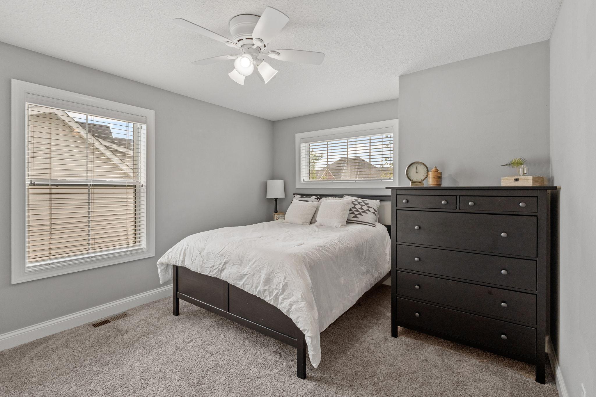 Bedroom with white bedding, dark wood furniture, gray walls, and two windows with blinds.