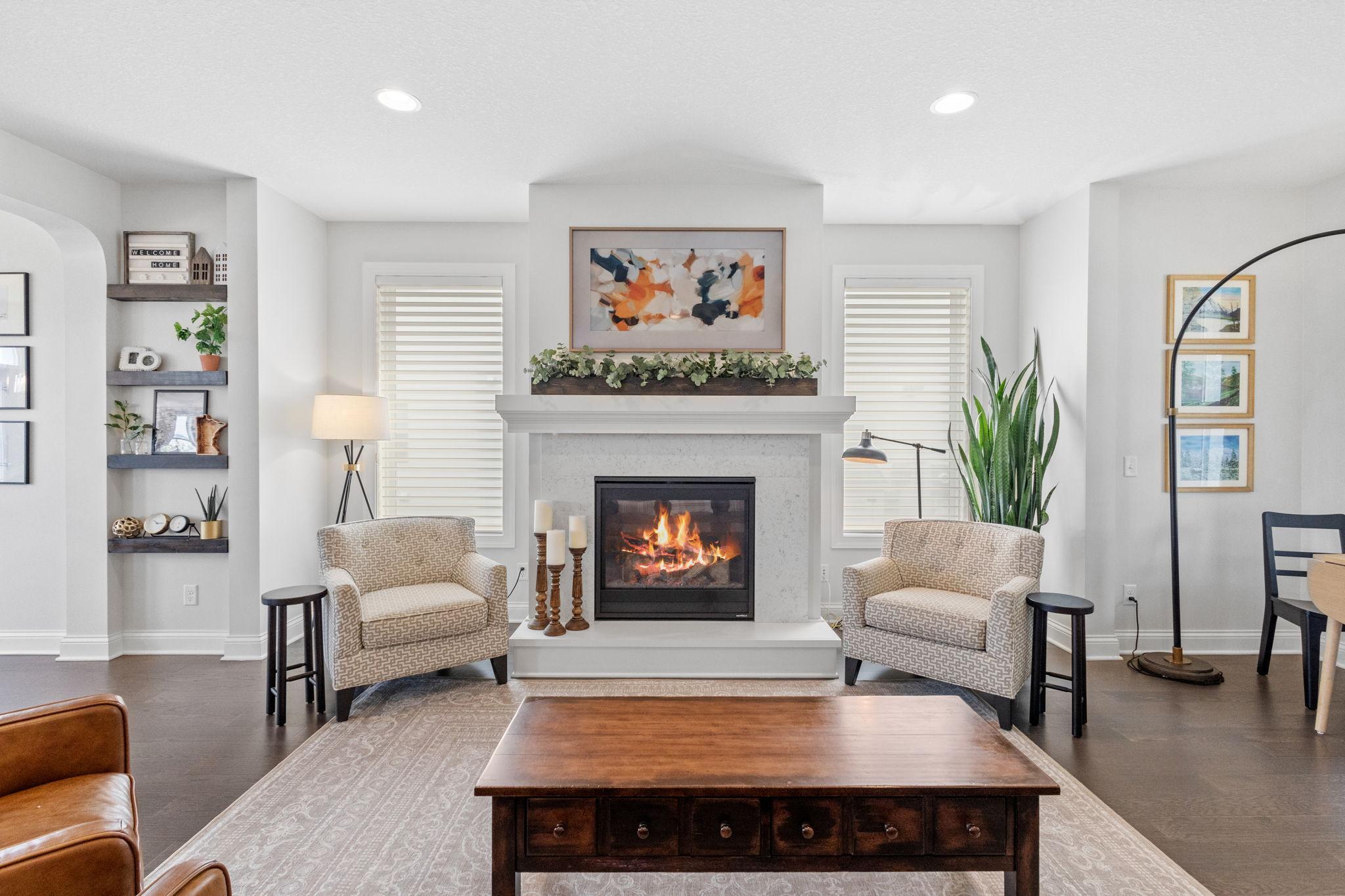 Staged living room with a lit fireplace, patterned armchairs, and built-in display shelves.