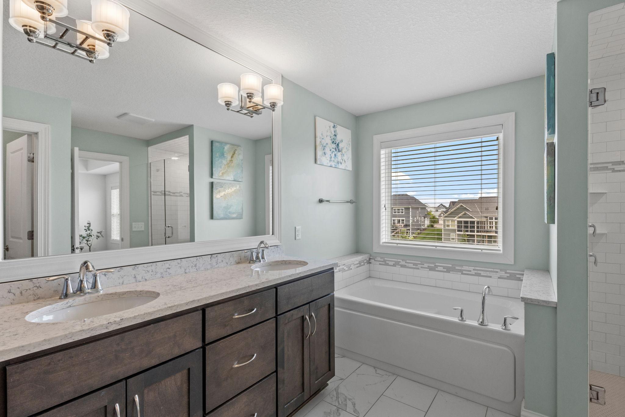 Bathroom with a dark double vanity, white tub, tiled shower, and window overlooking neighboring homes.