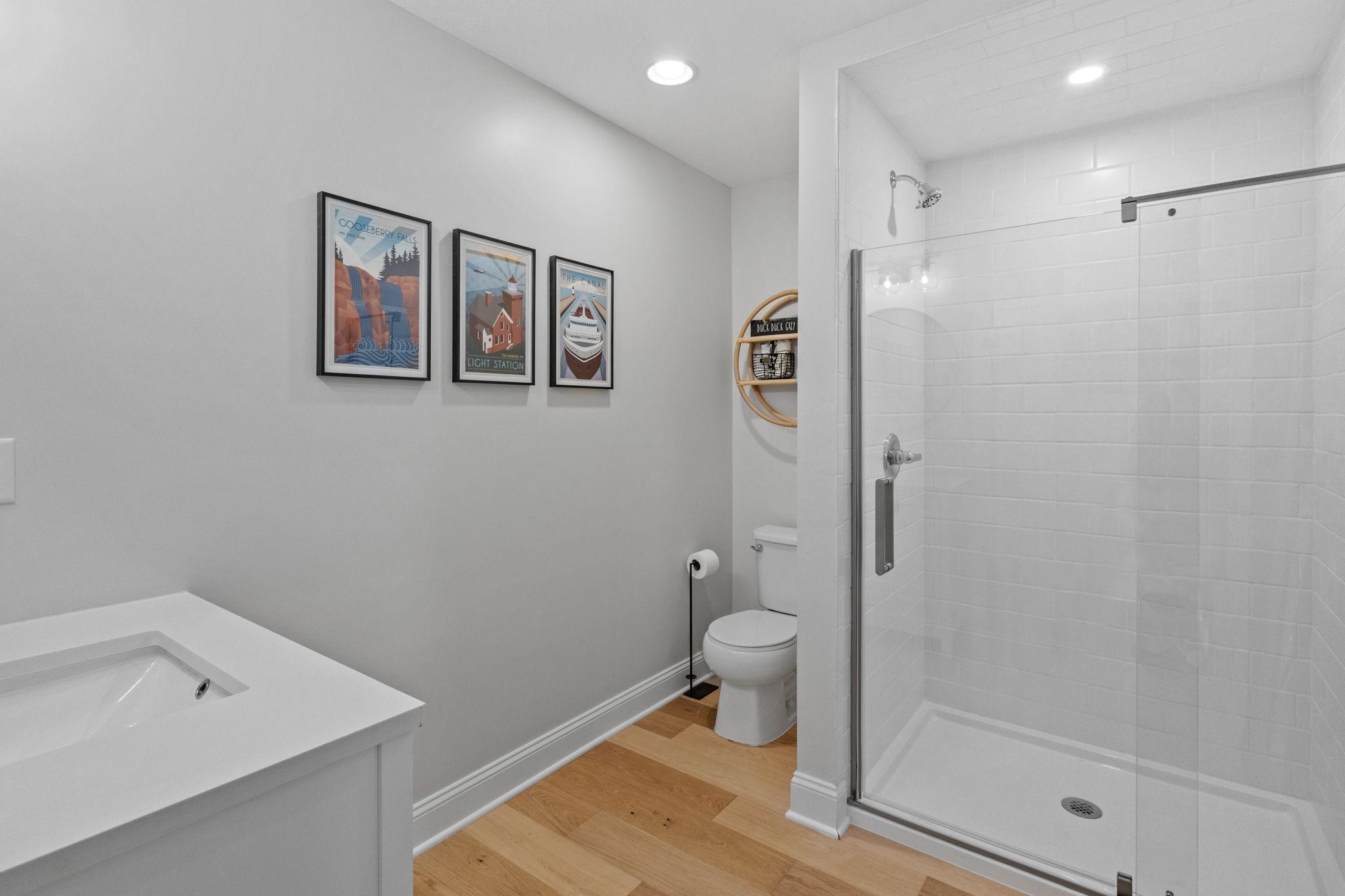 Bathroom with a glass-enclosed white-tile shower, white vanity, toilet, and warm wood flooring.