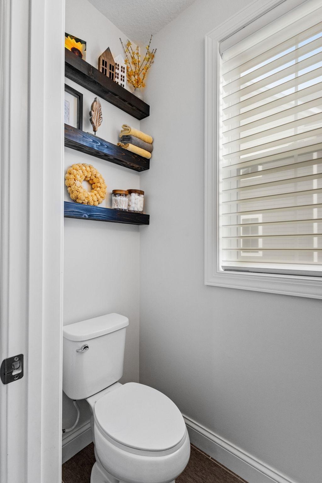 Powder room with a white toilet, dark floating shelves, and a window with blinds.
