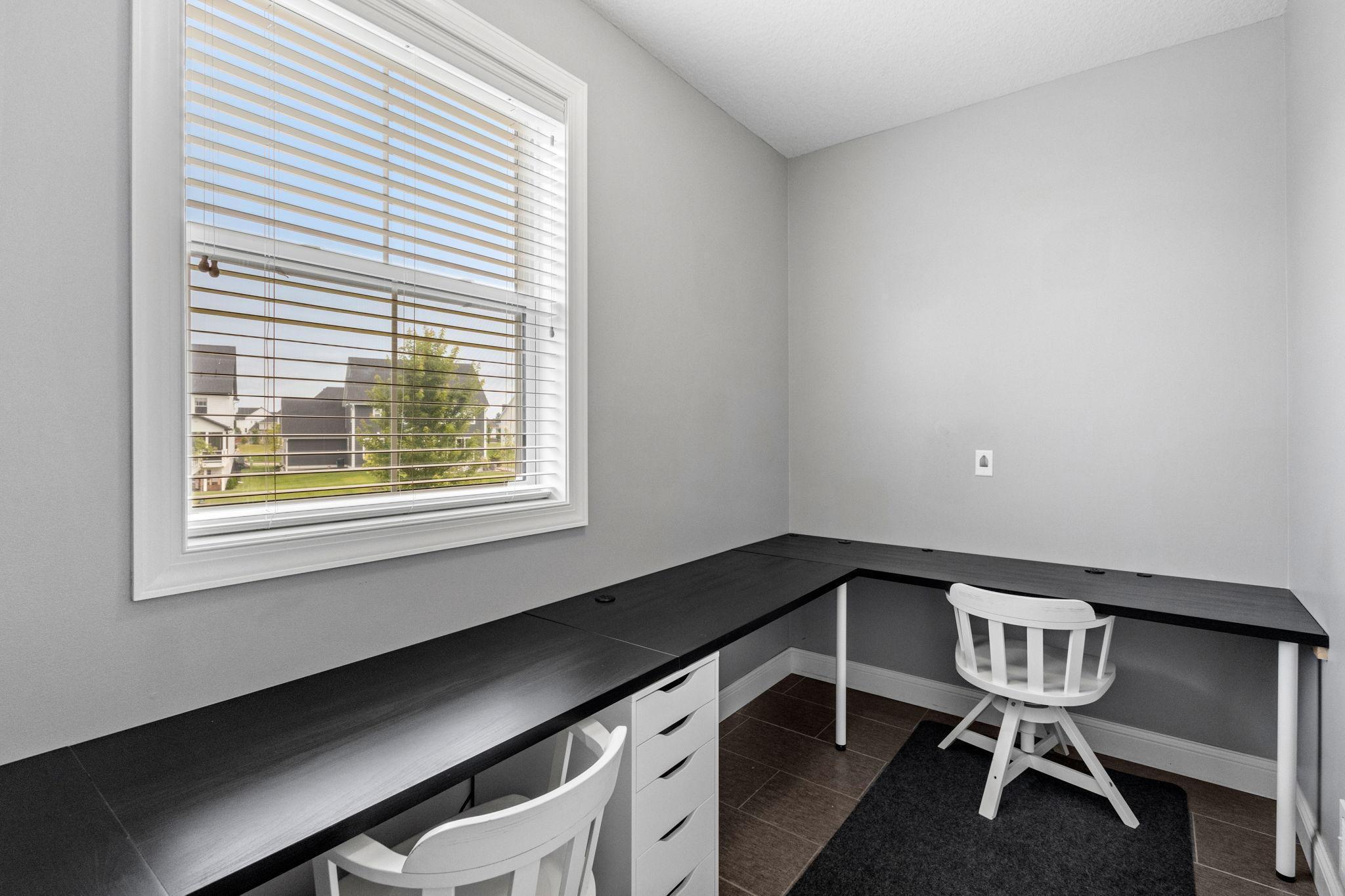 Home office with an L-shaped black desk, two white chairs, and a large window with blinds.
